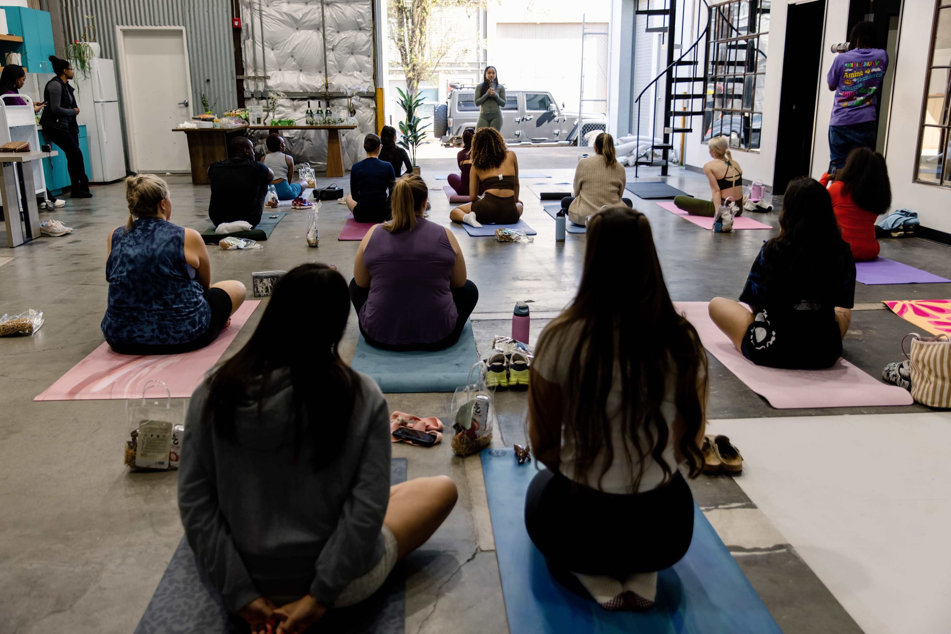 A group of people sits on yoga mats in a spacious indoor environment, listening to an instructor at the front.
