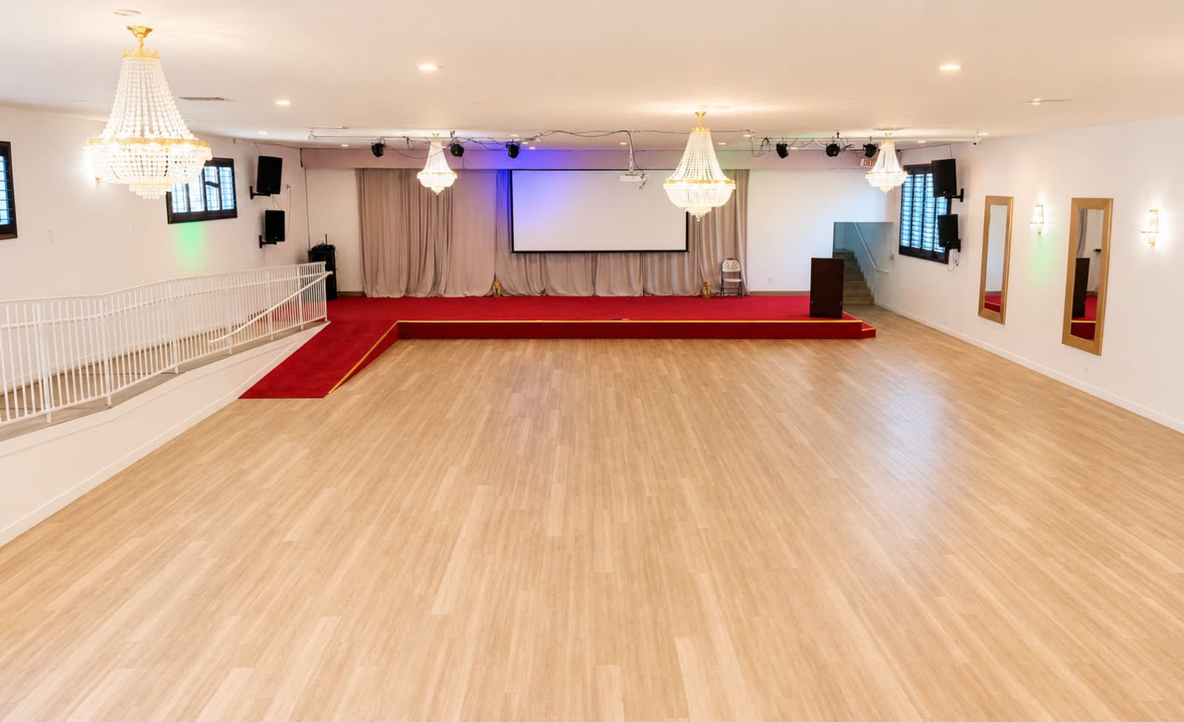 The image shows a spacious indoor venue with a wooden floor, stage area, and chandeliers, set for an event.
