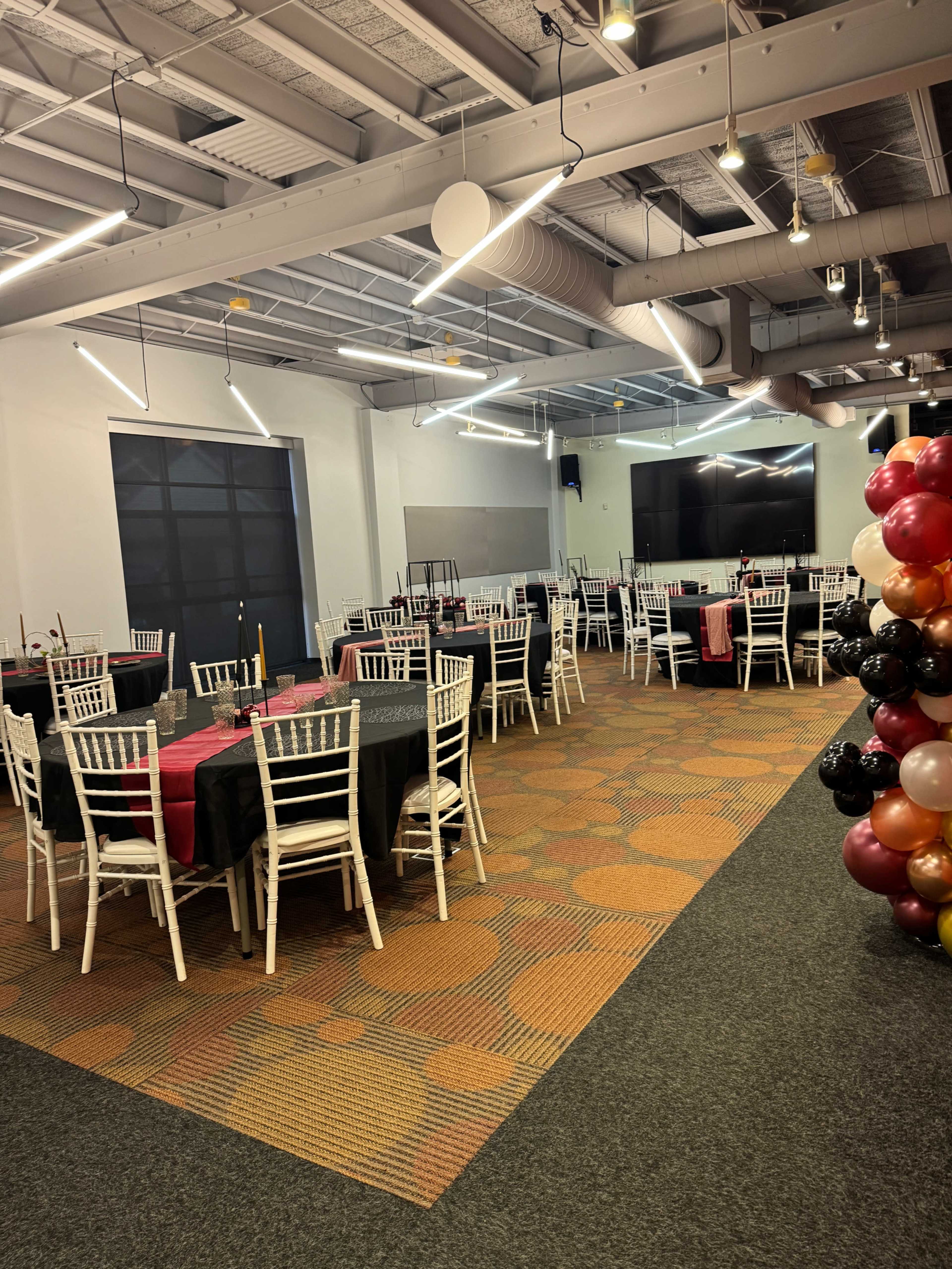 A banquet hall with several tables covered in black and red tablecloths, arranged with white chairs, and decorative balloons in the corner.