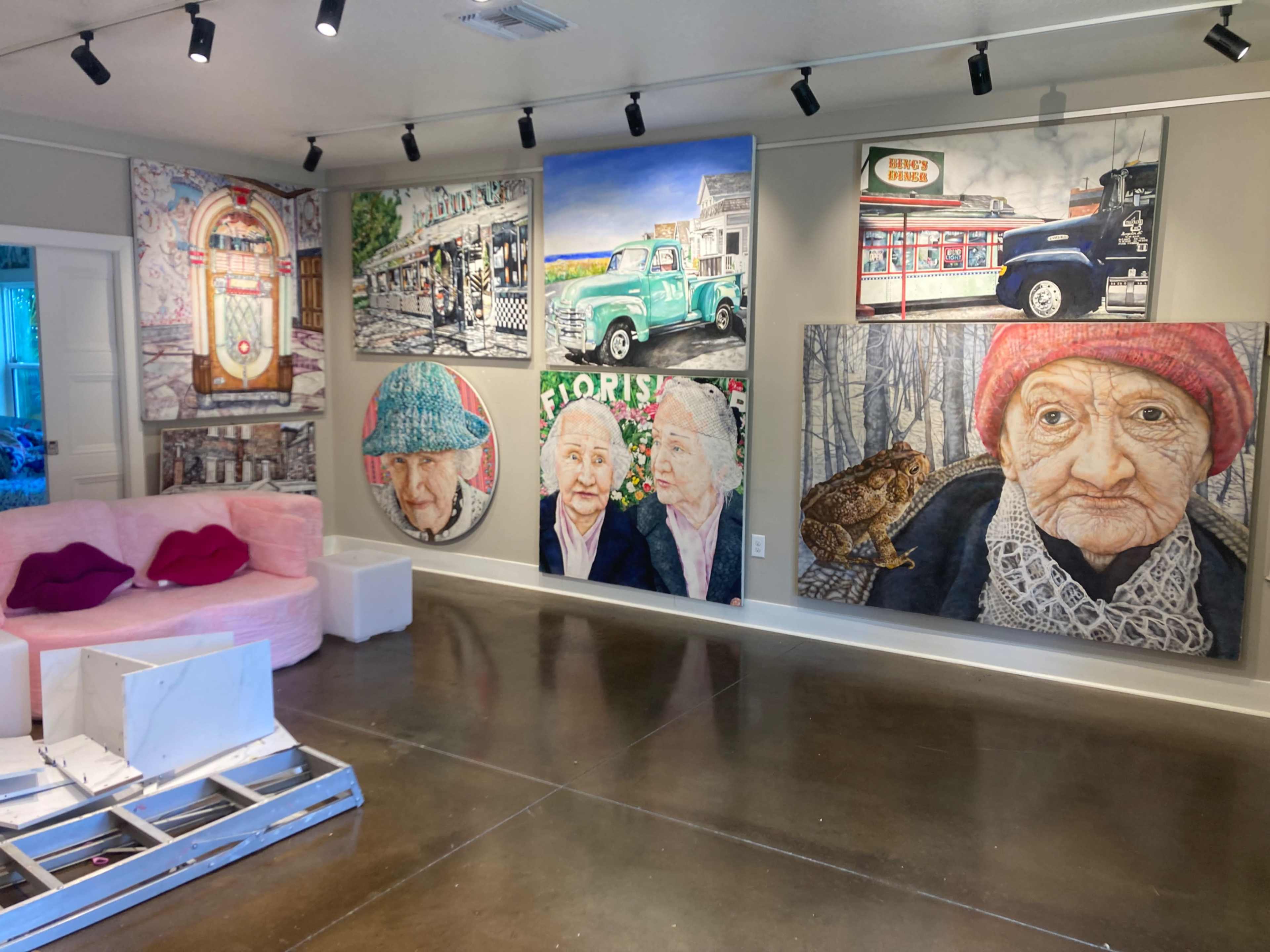 The image shows an art gallery featuring large portraits of elderly individuals on the walls, along with other colorful artworks and a pink sofa in the foreground.