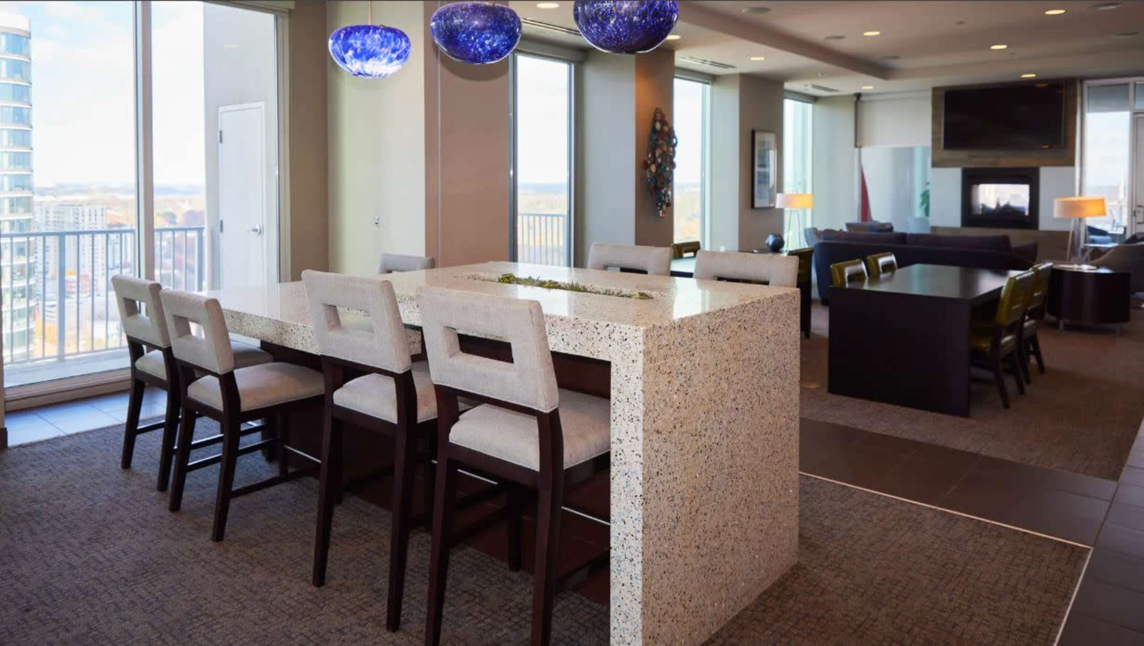 A modern kitchen and dining area features a large stone island with bar seating, complemented by pendant lighting and large windows offering a view of the outside.