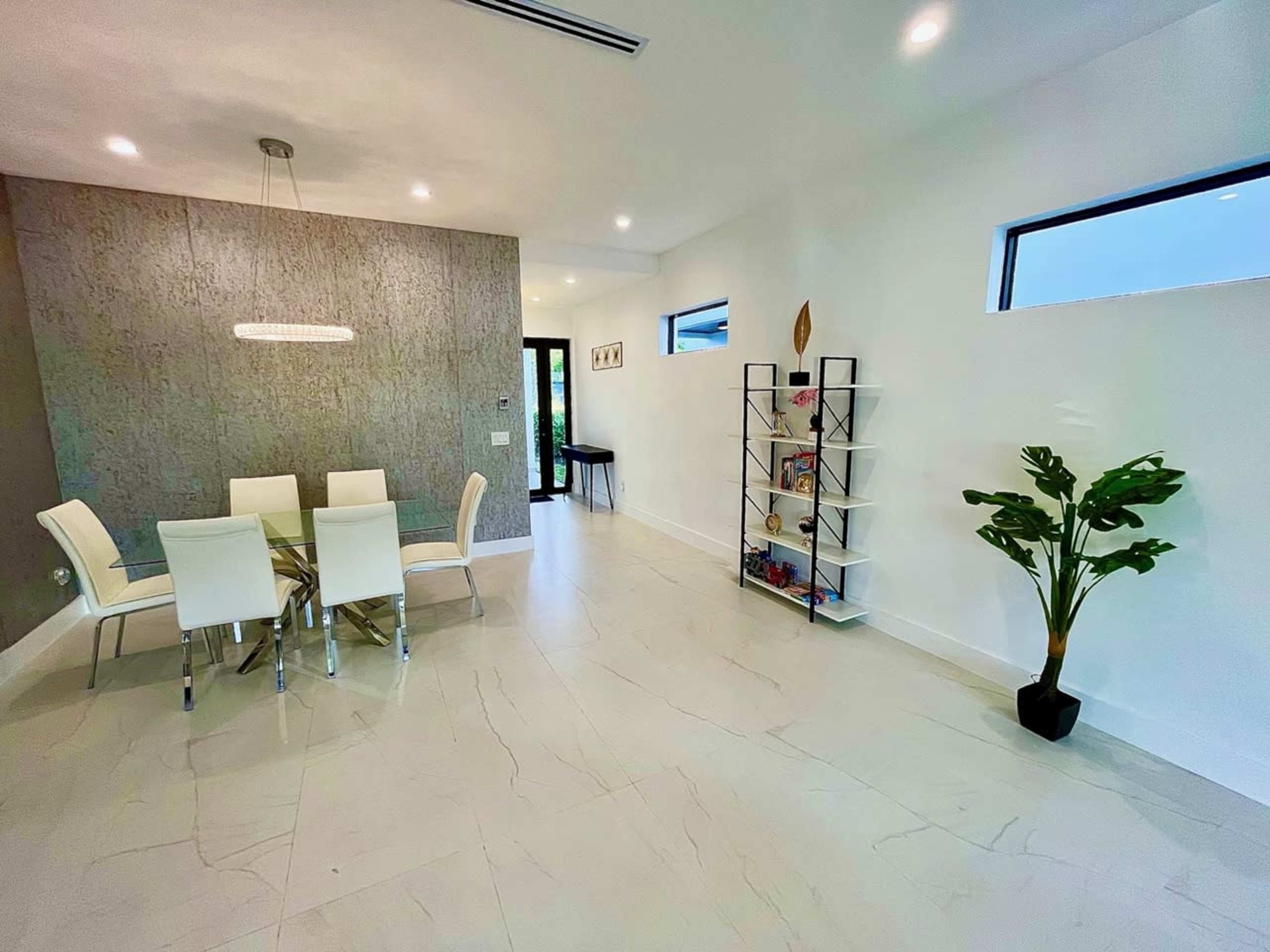 A modern dining area features a glass table with four white chairs, a decorative wall, a tall bookshelf filled with items, and a potted plant in the corner.