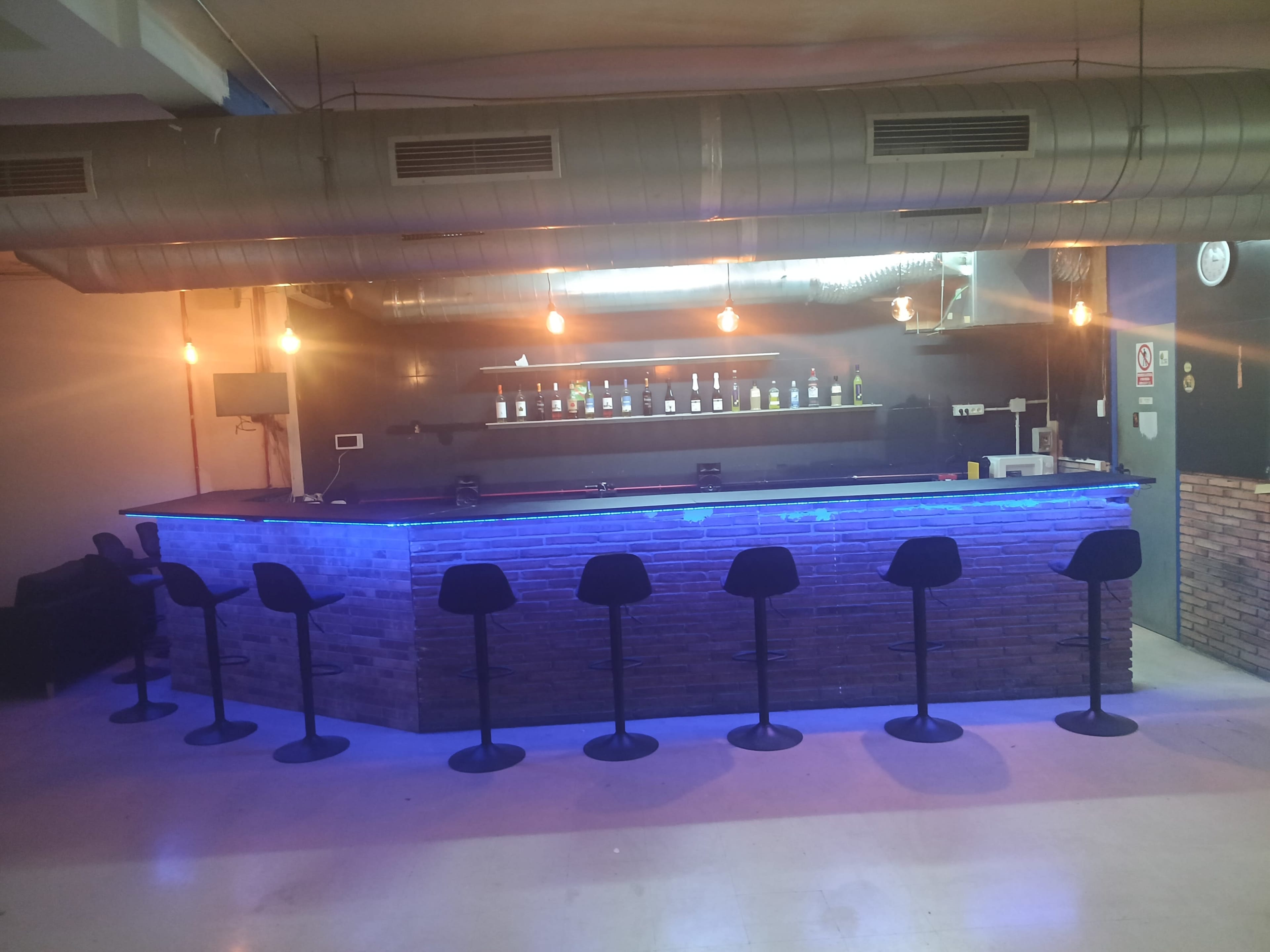 The image shows a bar area with a stone counter and several black stools, illuminated by overhead lights and blue LED accents.