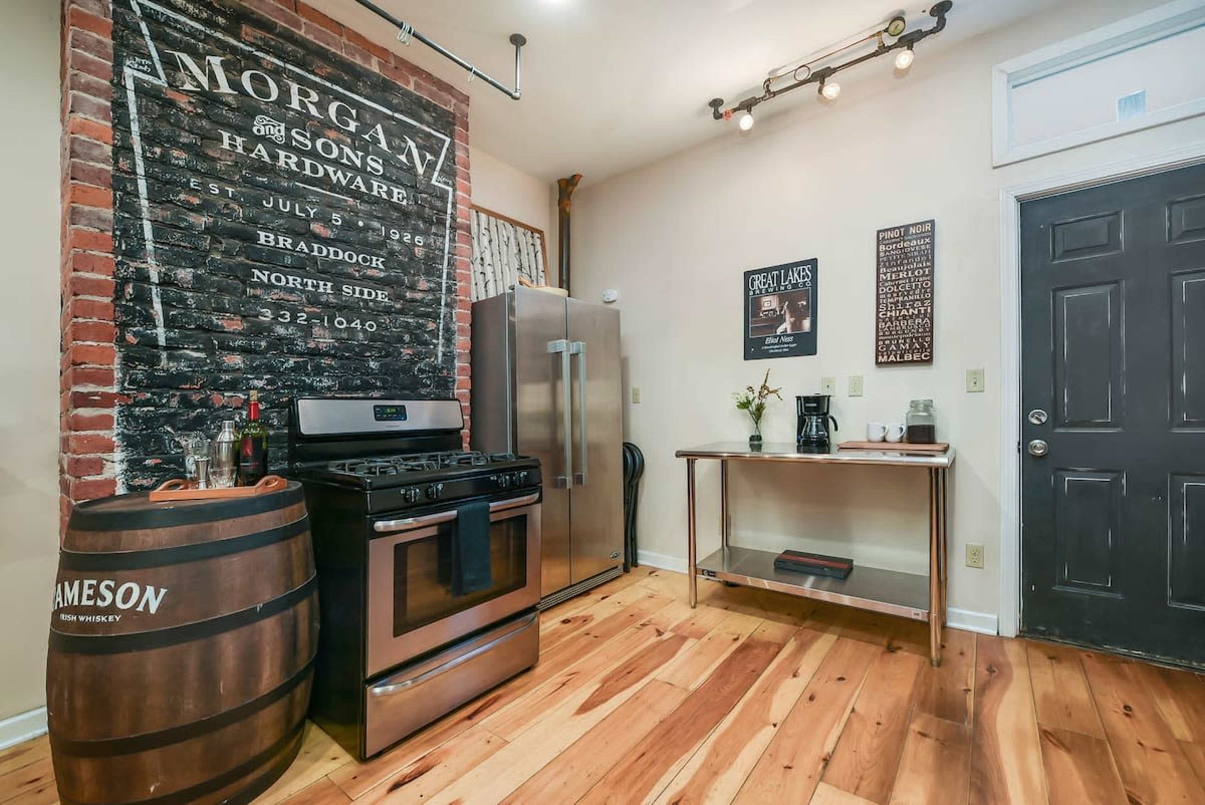 A kitchen features a stainless steel stove and fridge, exposed brick wall with a vintage sign, and wooden flooring.