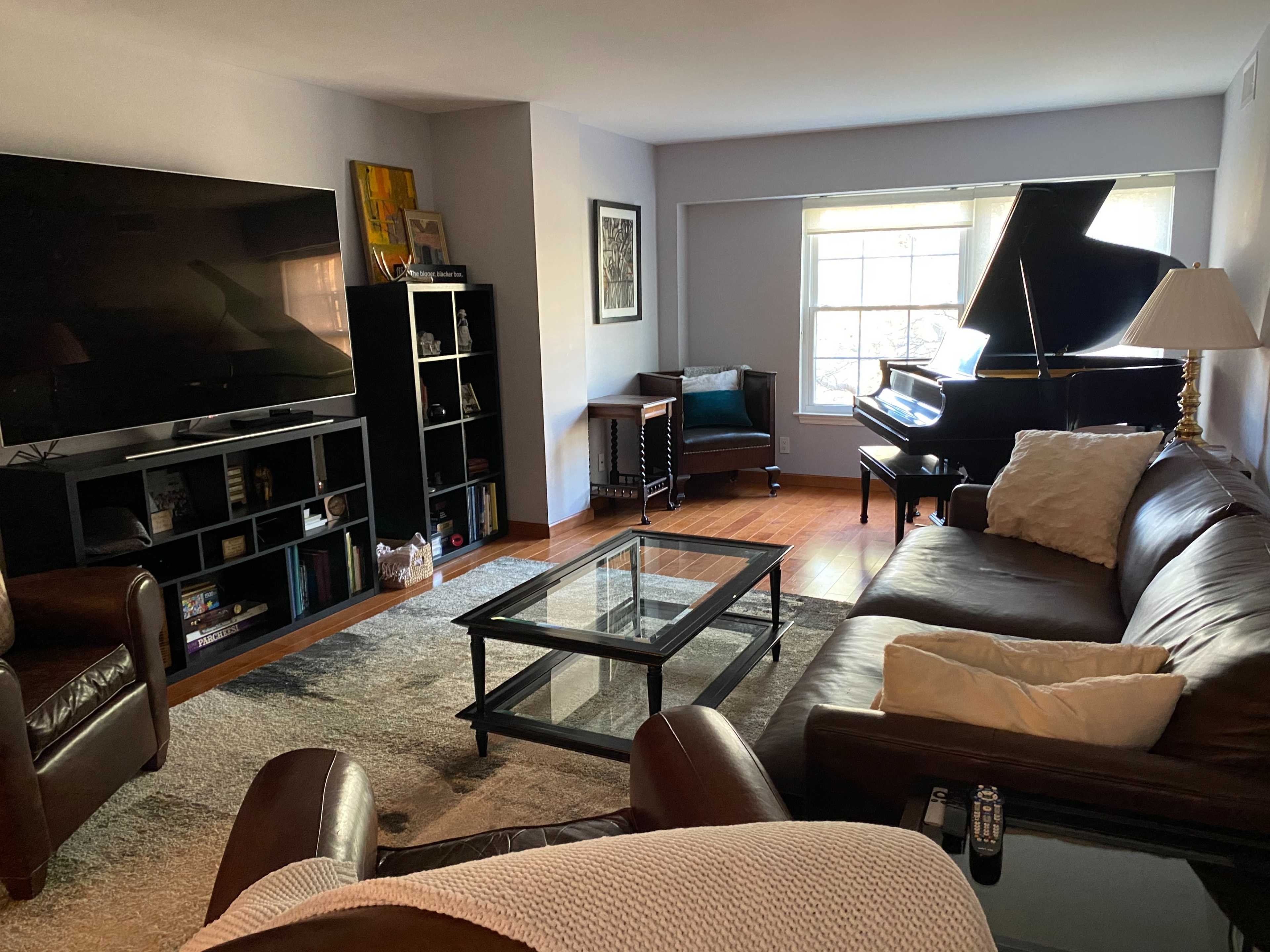 A living room features a large television, a glass coffee table, a leather sofa, a grand piano, and a cozy armchair by a window.