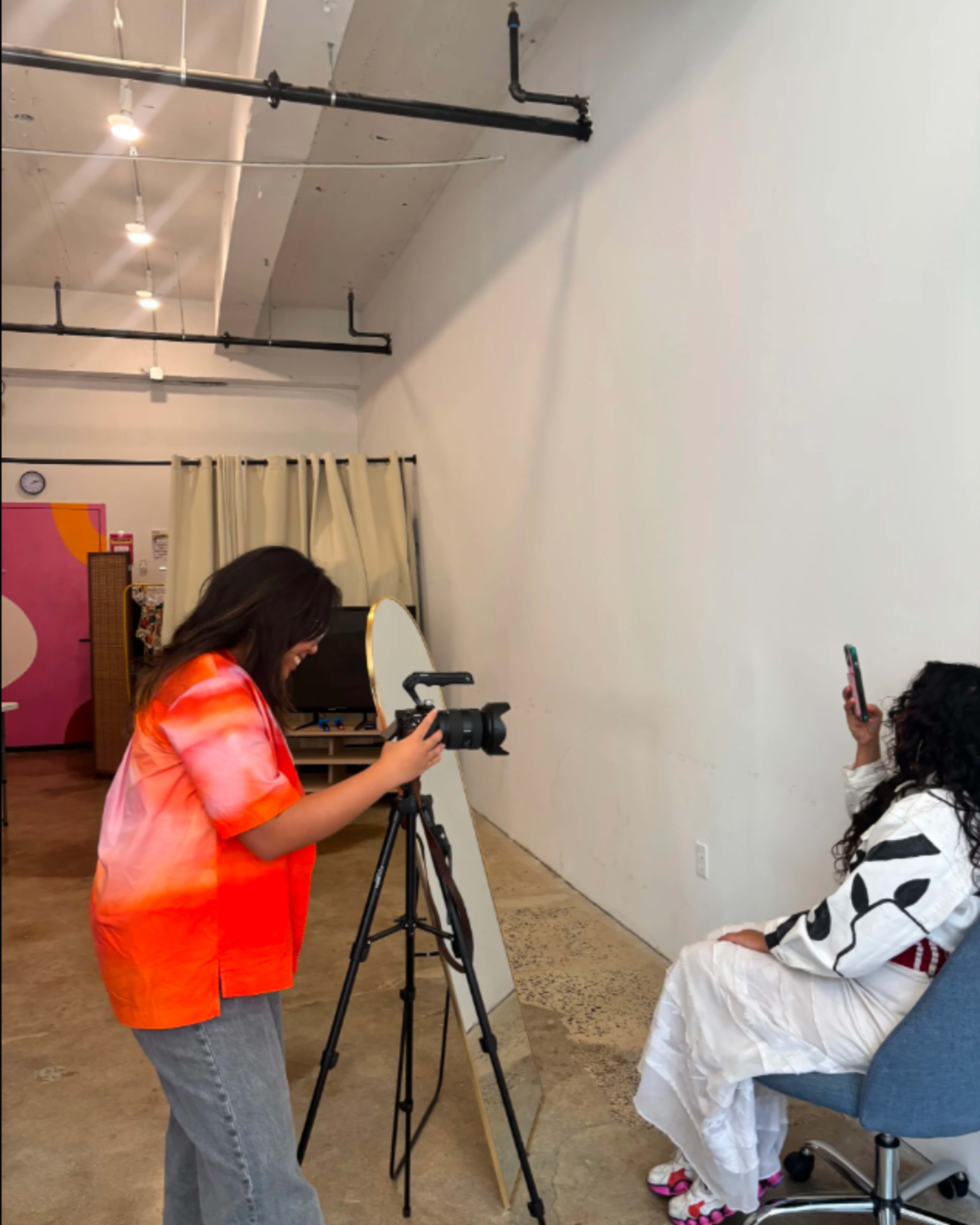A photographer sets up a camera on a tripod while another person poses with a phone in front of a blank wall.