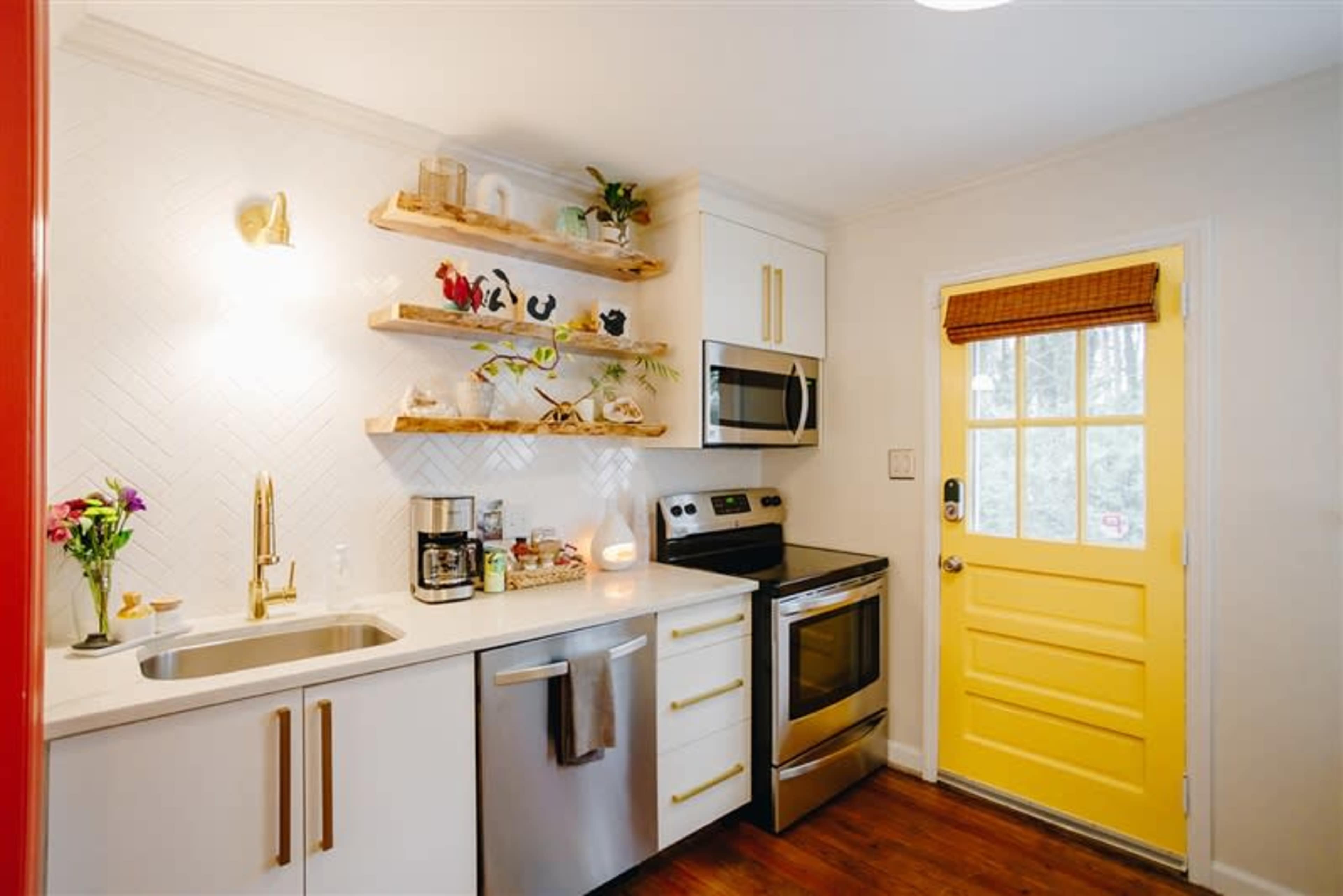 A small kitchen features open wooden shelves, stainless steel appliances, and a bright yellow door.