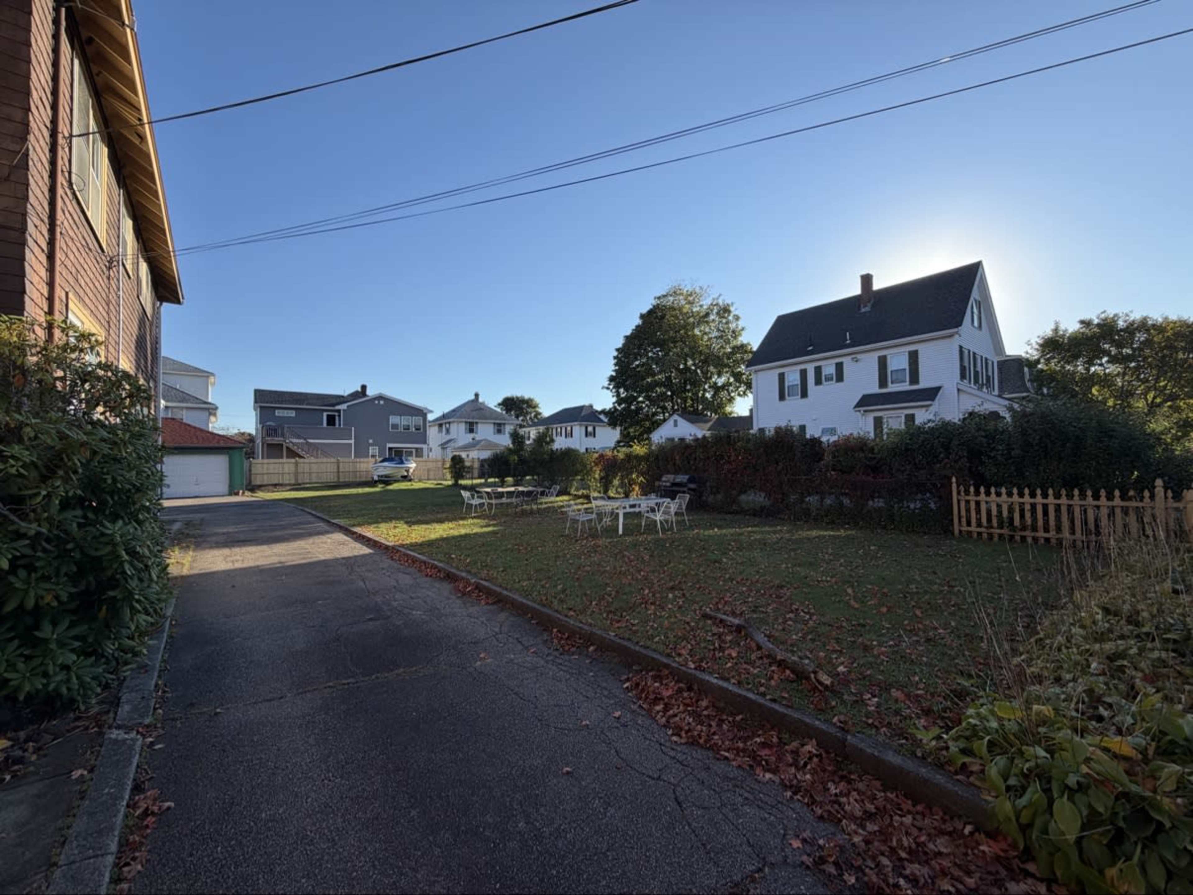 The image shows a residential area with a paved driveway, a lawn with scattered leaves, and several houses in the background under a clear blue sky.