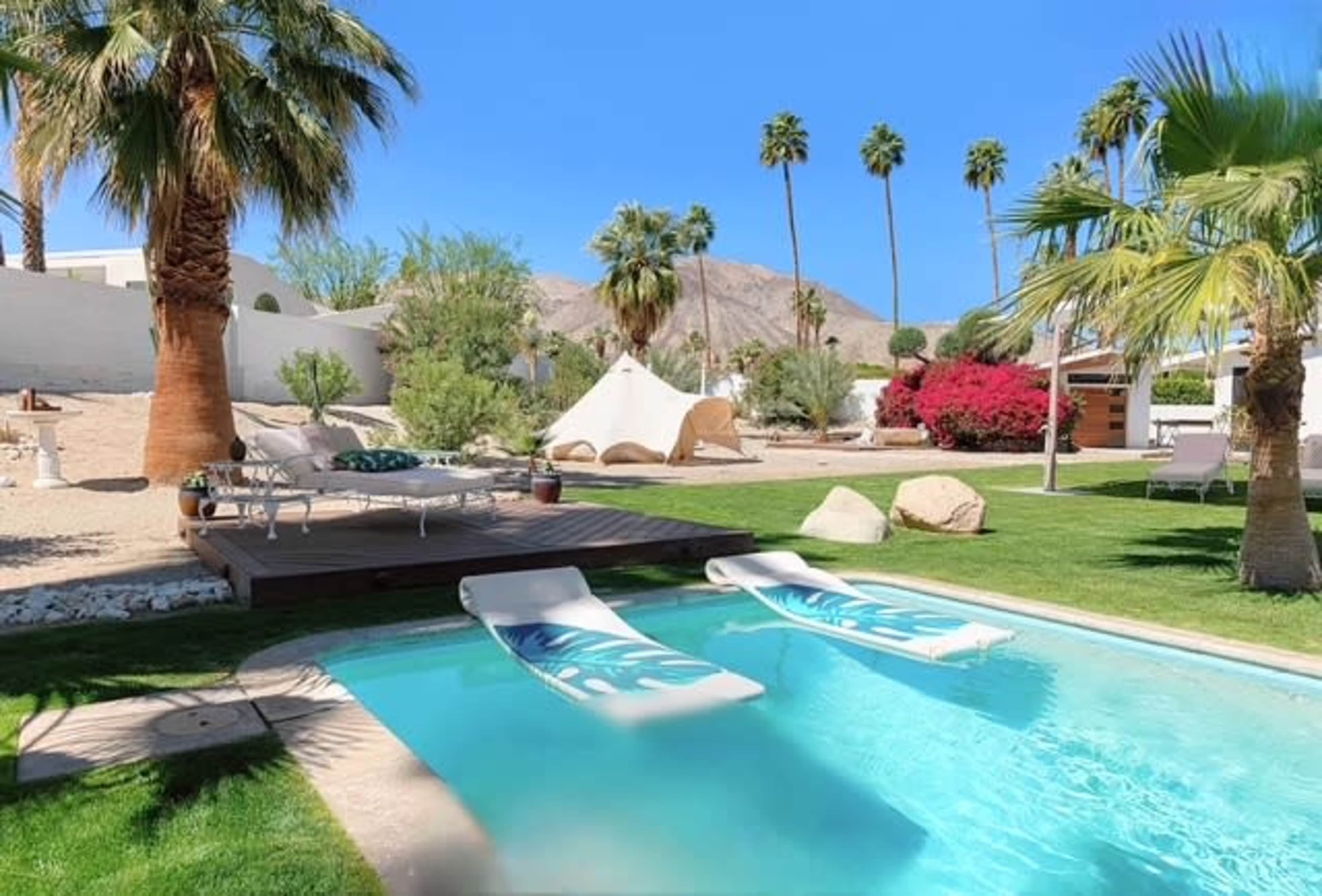 The image shows a backyard with a swimming pool, two pool loungers, palm trees, and a tent-like structure in a desert landscape.
