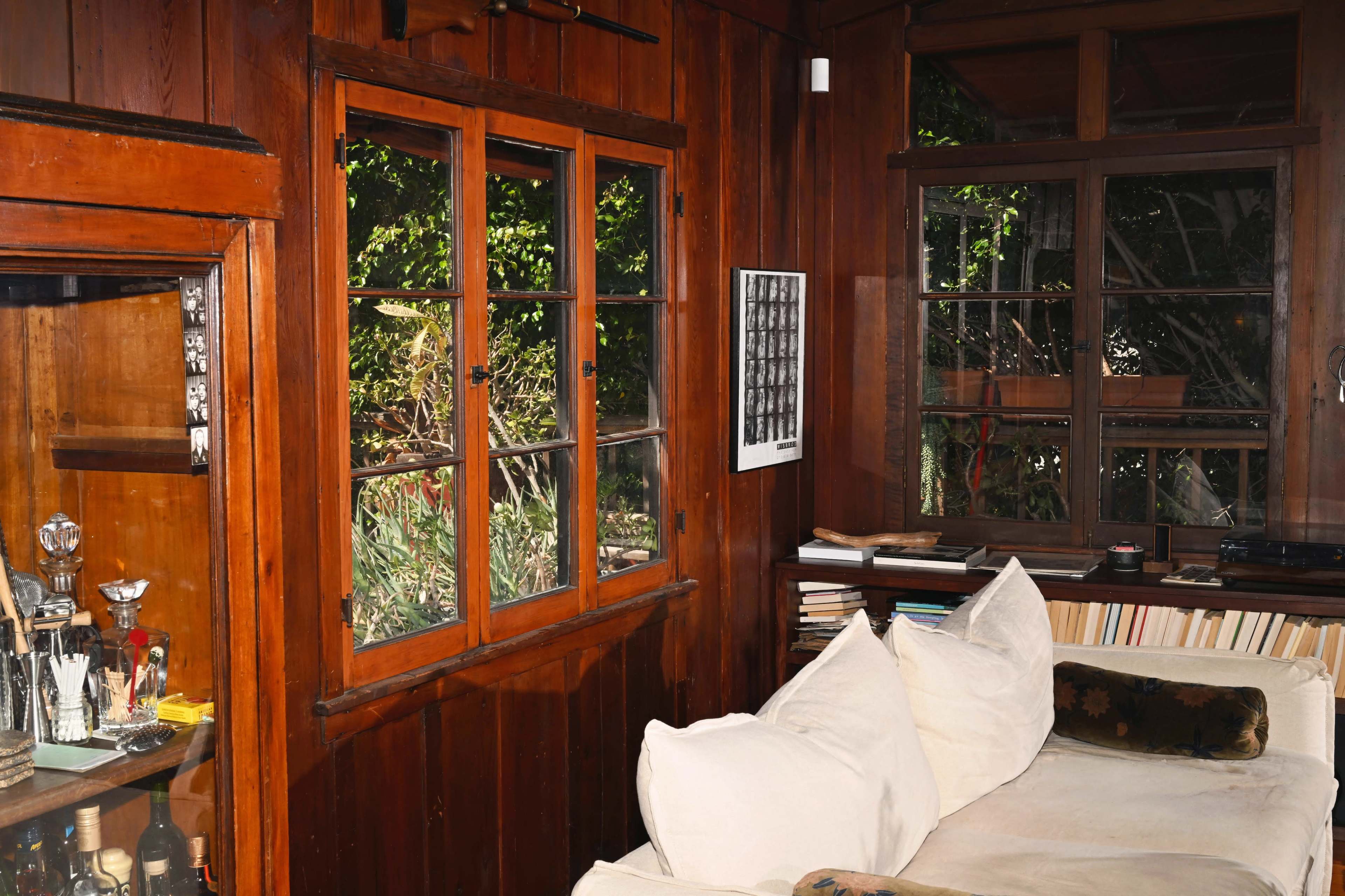 The image shows a wooden room with two windows, a couch adorned with white pillows, and a bookshelf against the wall.