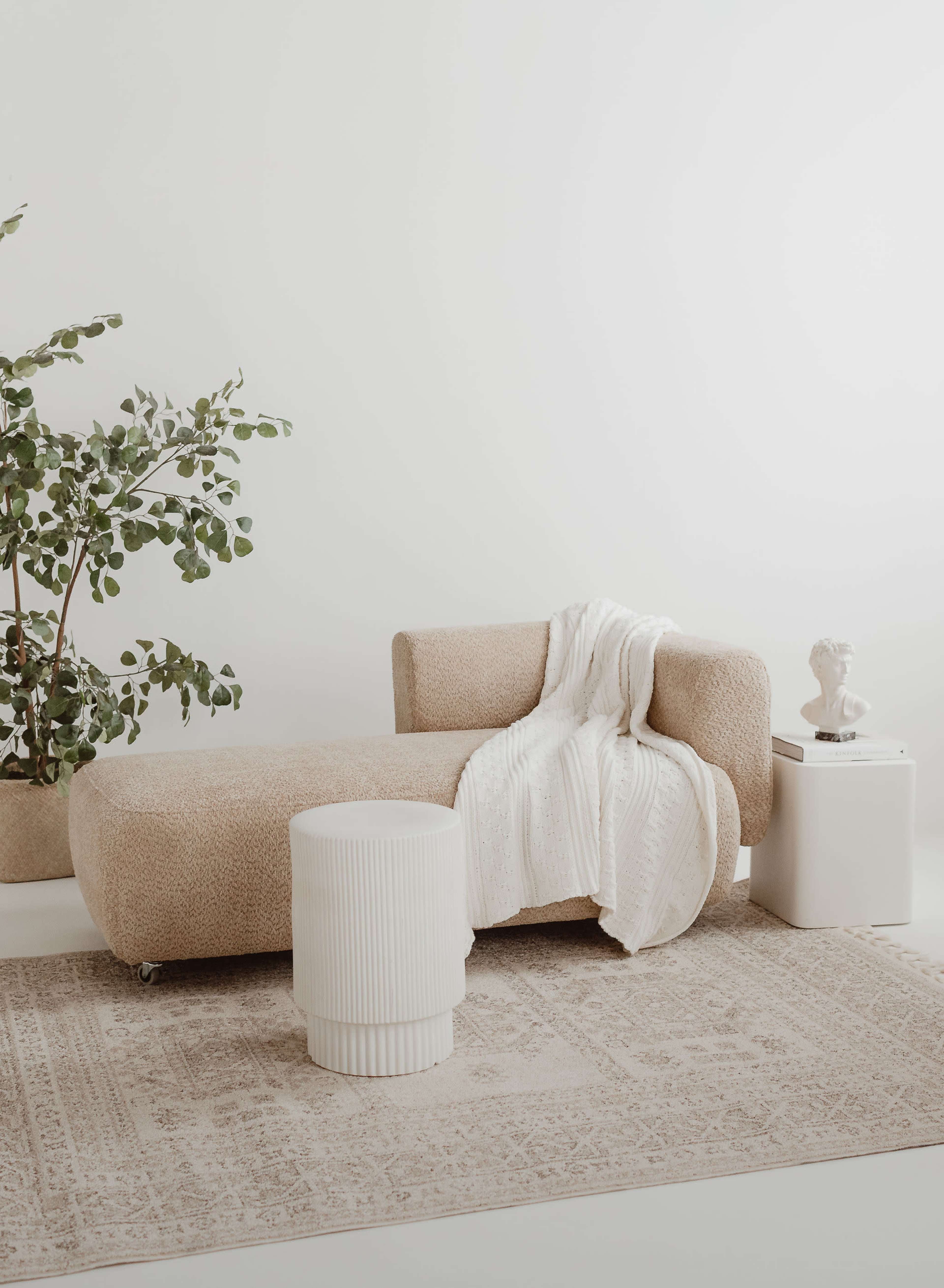 The image features a minimalist living space with a beige textured chaise lounge, a white blanket draped over it, a small round white side table, a decorative statue, and a potted green plant.