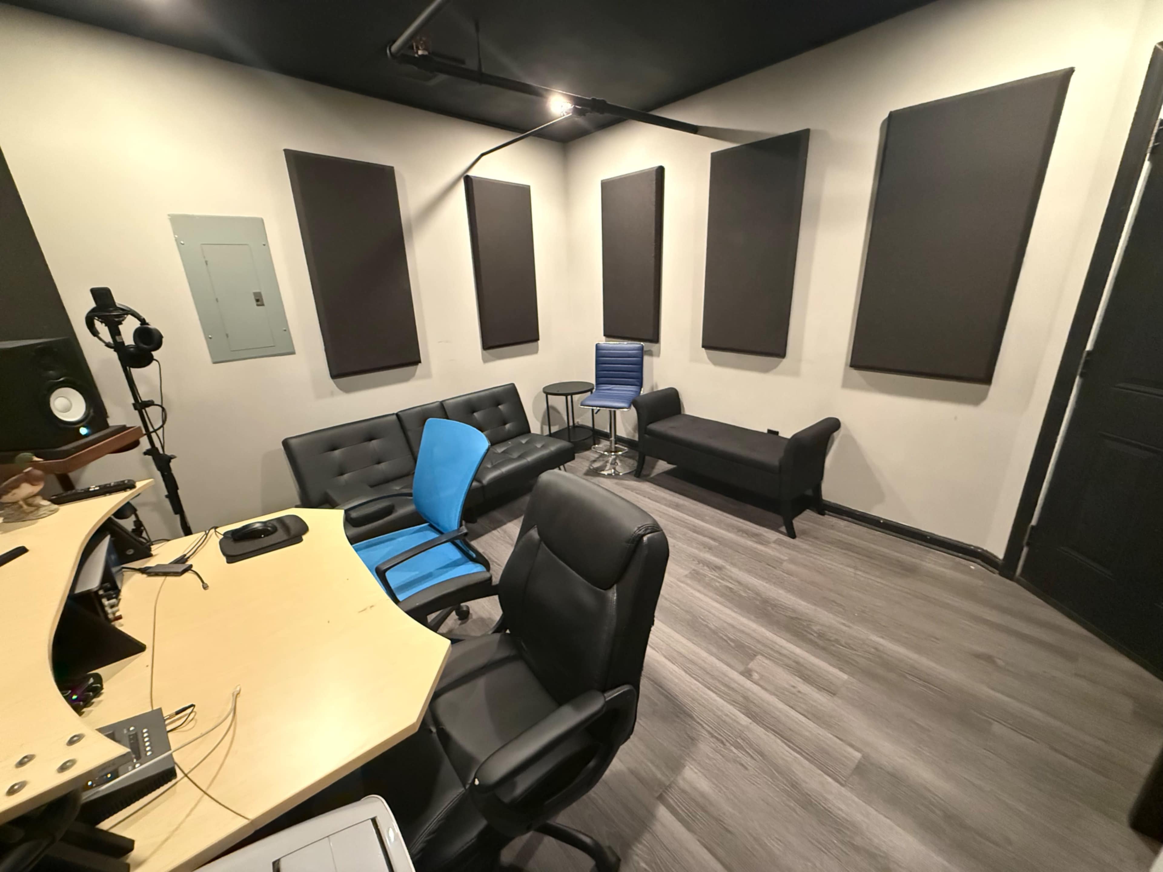 The image shows a soundproof recording studio with black acoustic panels on the walls, a desk equipped with audio equipment, and various seating options including a blue chair and a black sofa.