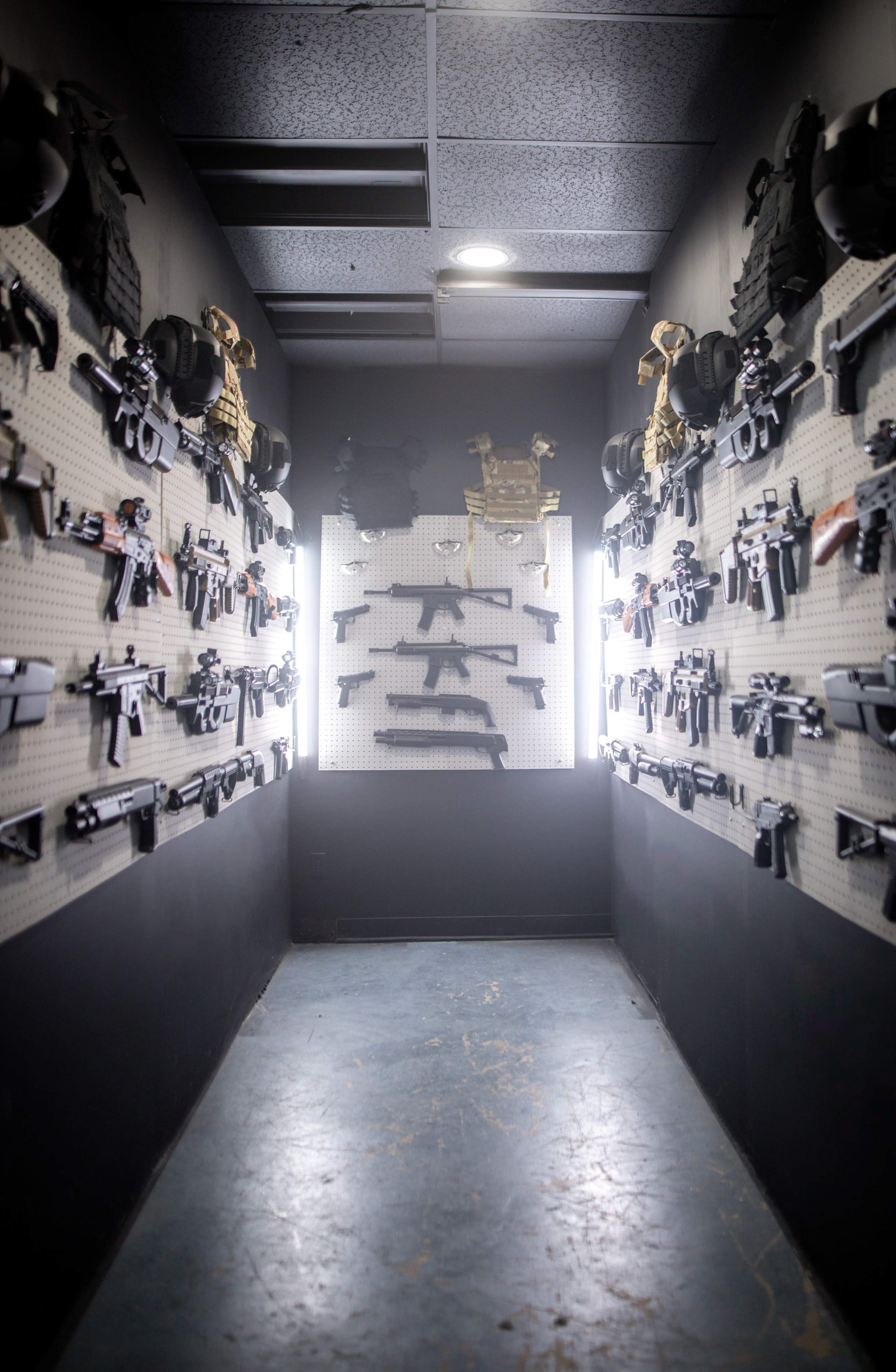 The image shows a narrow corridor lined with various firearms displayed on walls, with a spotlight illuminating a central arrangement of guns.