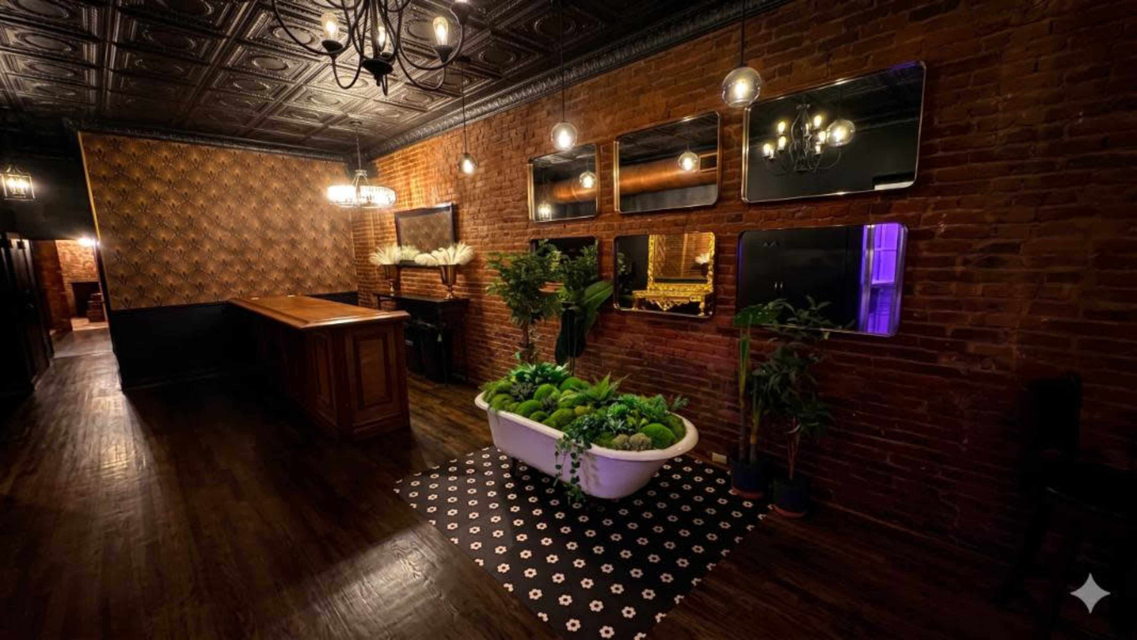 The image shows a stylish interior featuring exposed brick walls, a wooden bar, decorative mirrors, and a bathtub filled with greenery on a patterned rug.