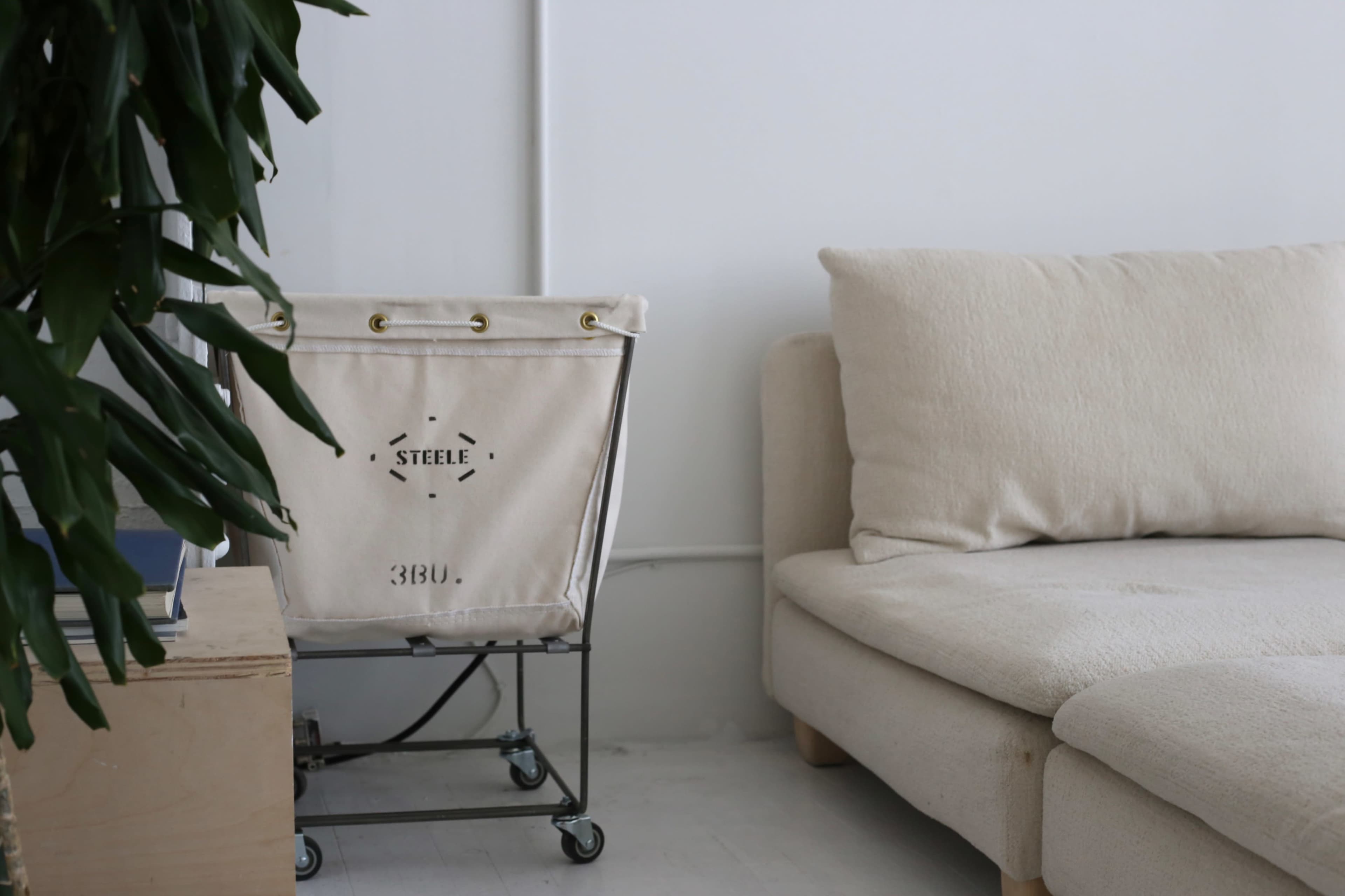 A minimalist living space features a light-colored sofa next to a wheeled laundry basket and a potted plant against a white wall.