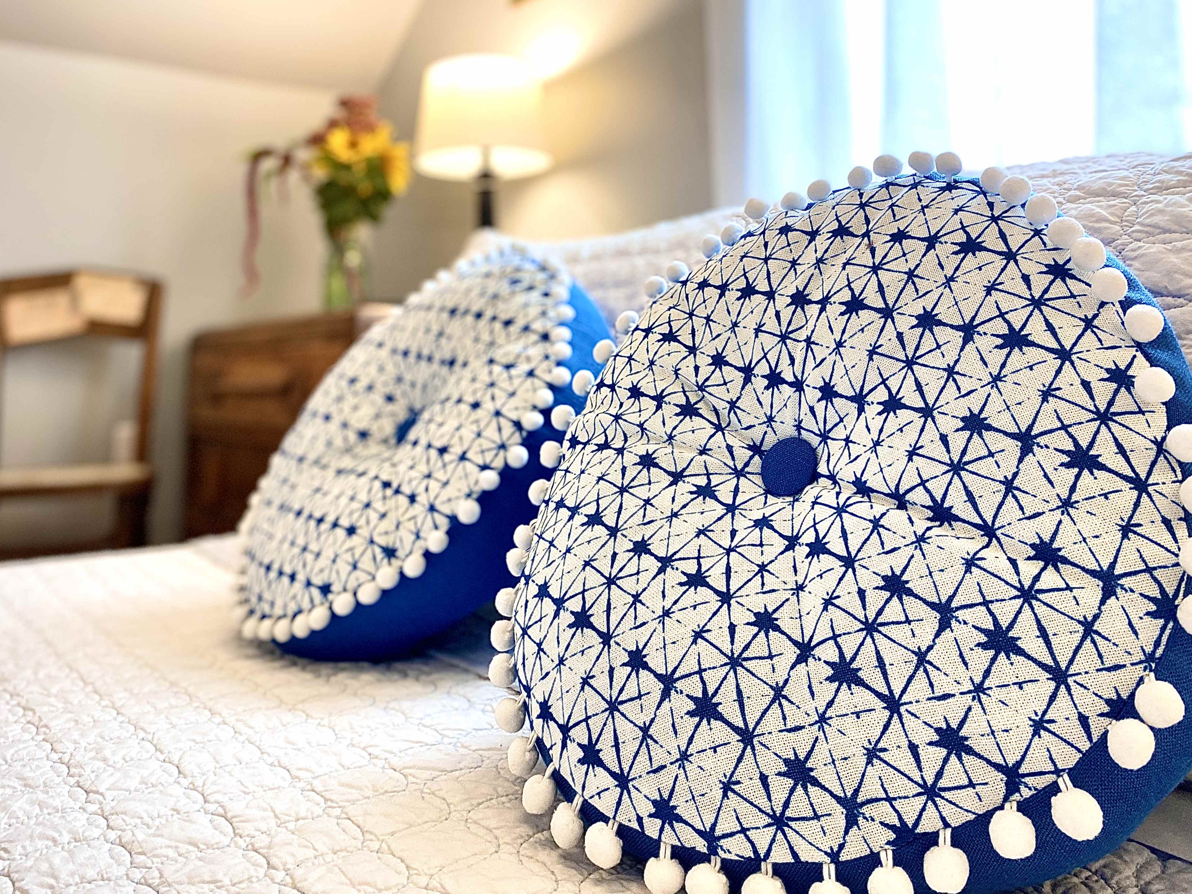 Two round blue and white decorative pillows with pom-pom edges rest on a neatly made bed in a softly lit room.