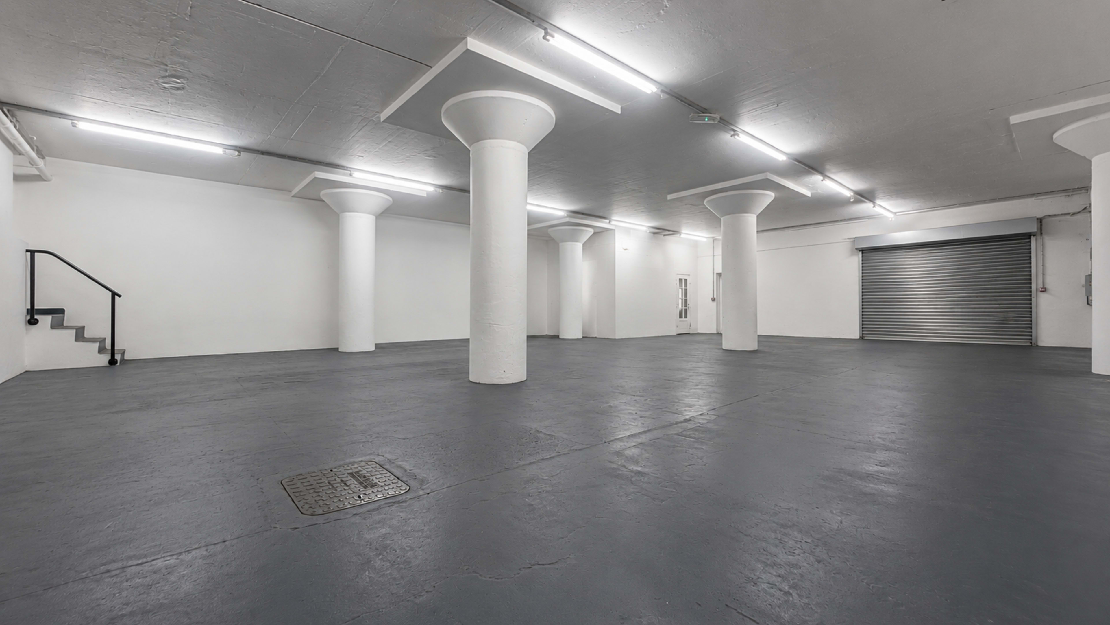 The image shows a large, empty industrial space with concrete flooring and several white columns, illuminated by fluorescent lights.