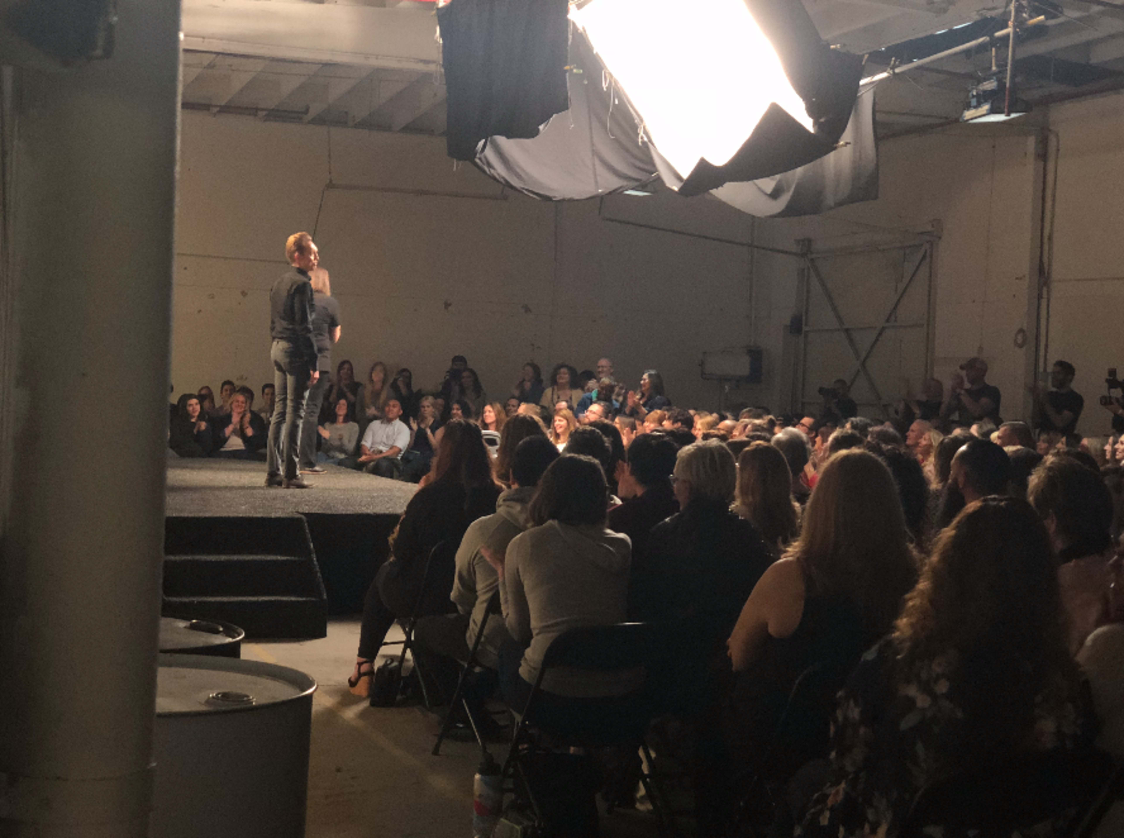 A person stands on a stage in front of a large audience in a warehouse-like space.