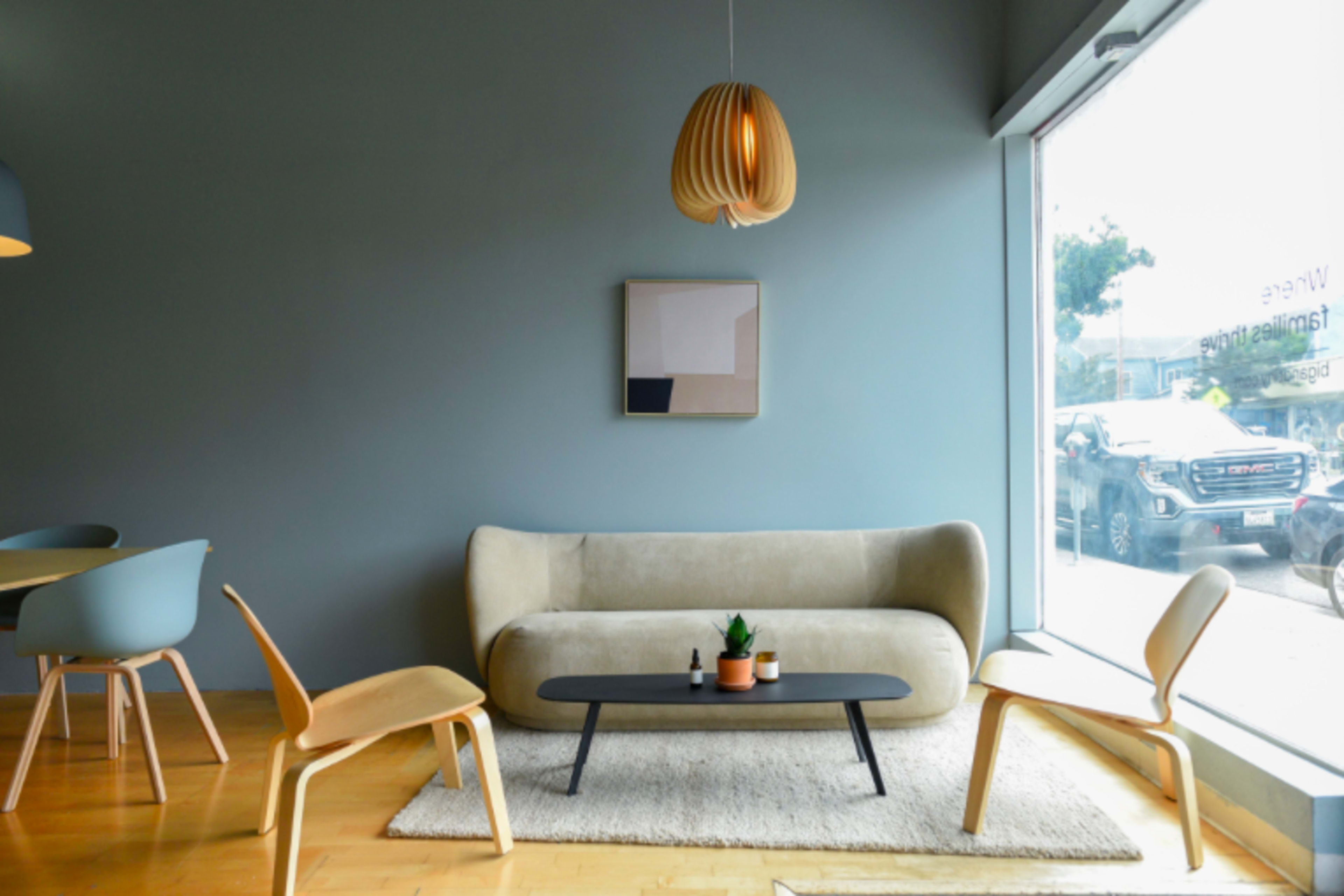 A minimalist living room features a curved beige sofa, a low black coffee table with a plant, and wooden chairs, bathed in natural light from a large window.