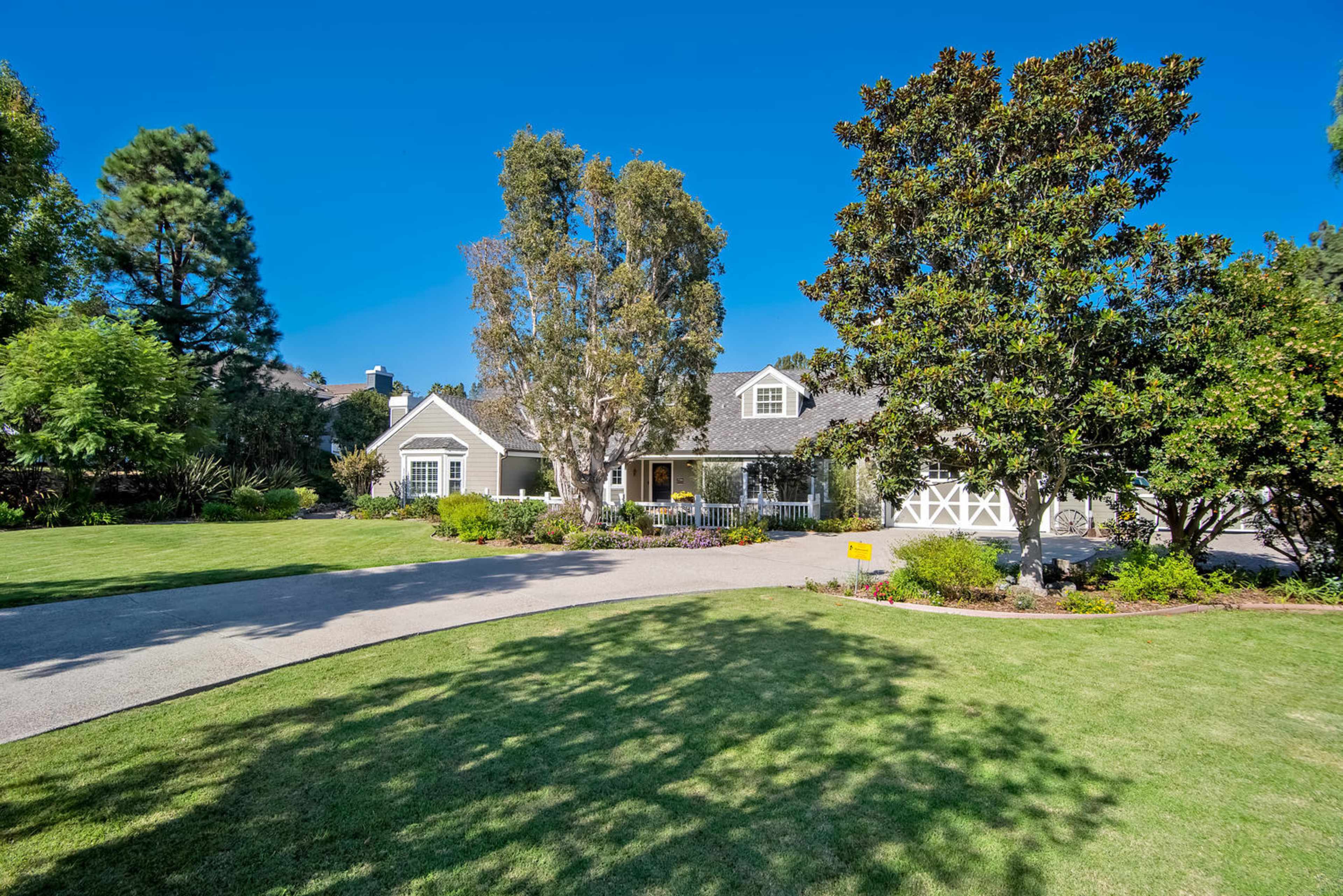 The image shows a single-story house with a large front yard and driveway, framed by several trees and shrubs under a clear blue sky.