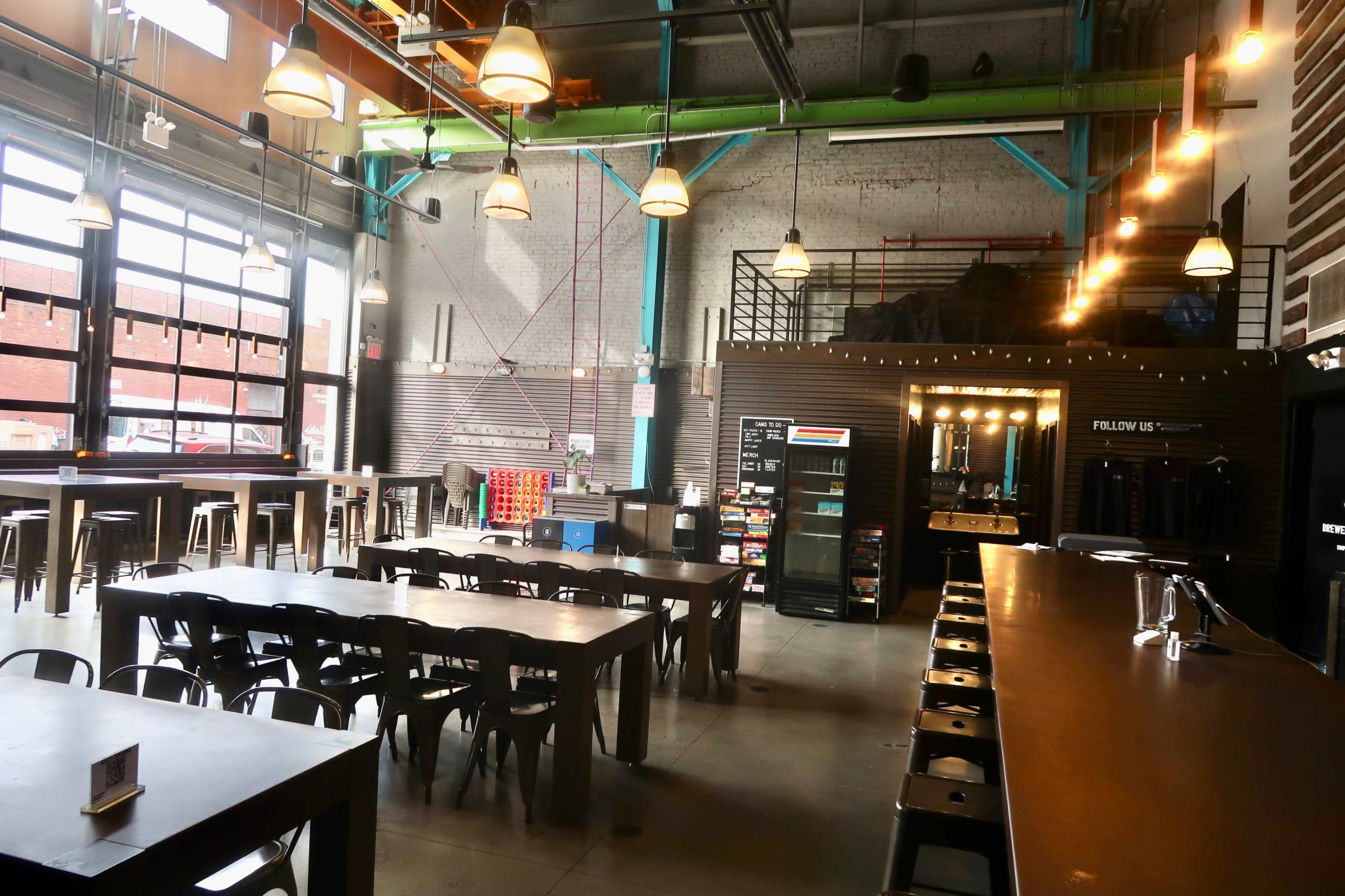 The image shows a spacious indoor venue with high ceilings, featuring long tables, bar stools, and a well-lit bar area with industrial decor elements.