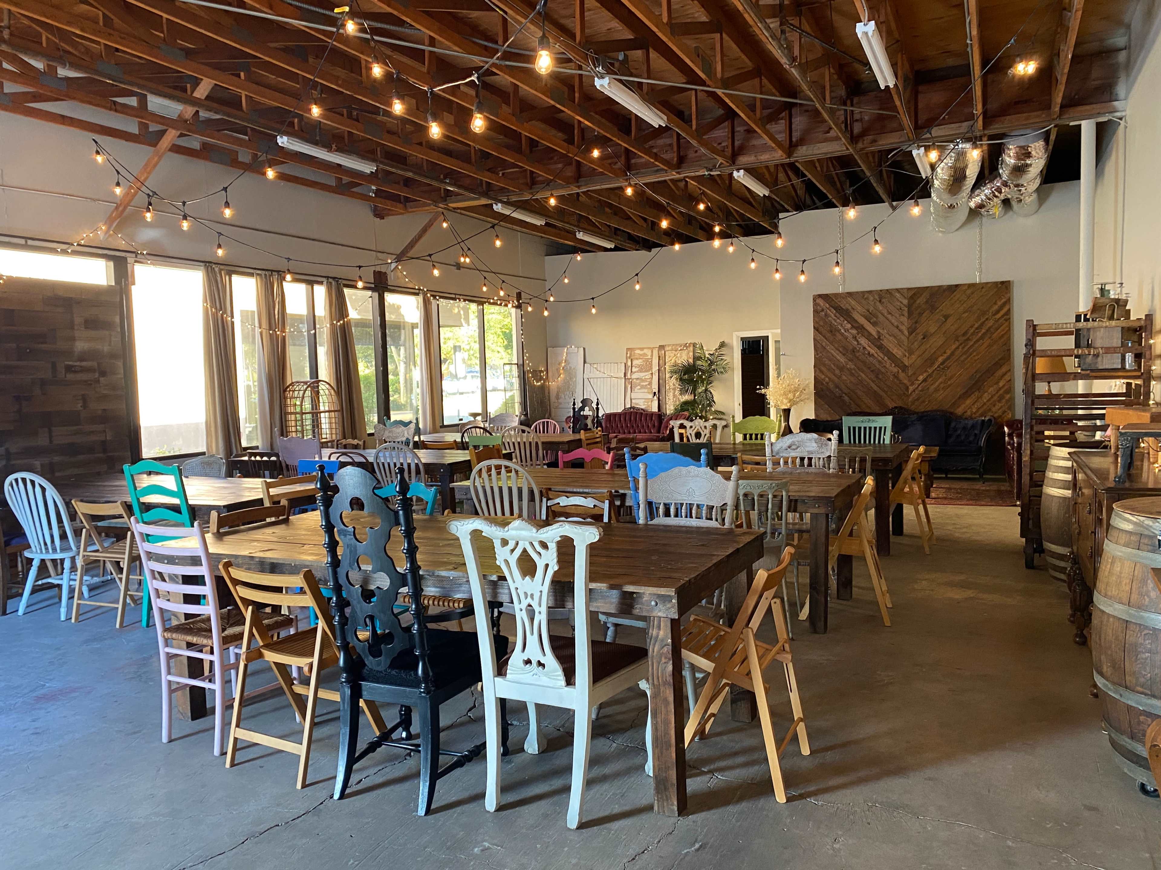 A spacious indoor gathering area filled with various wooden tables and colorful chairs, featuring string lights overhead and large windows letting in natural light.