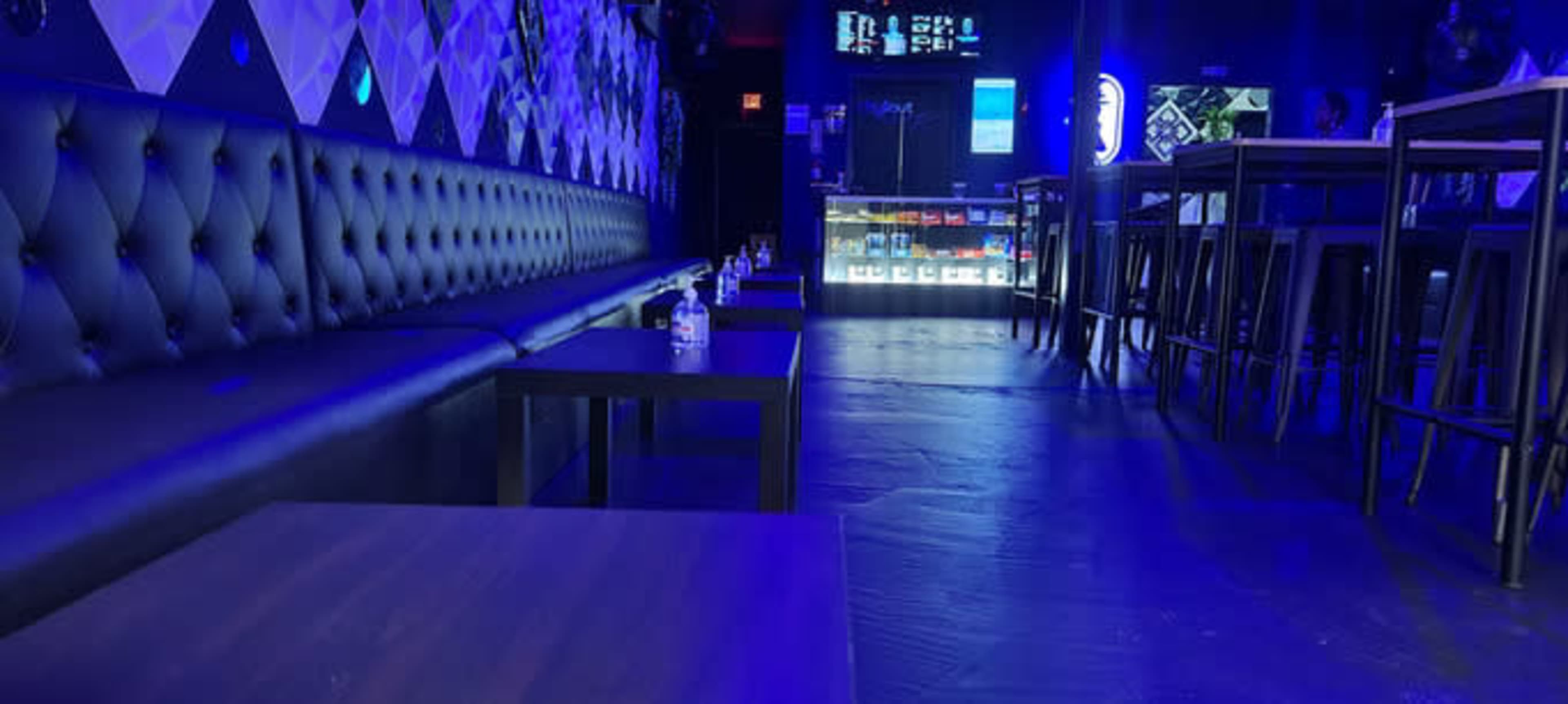 The image shows a modern lounge area with black leather seating, dark tables, and illuminated shelves displaying various products in the background.