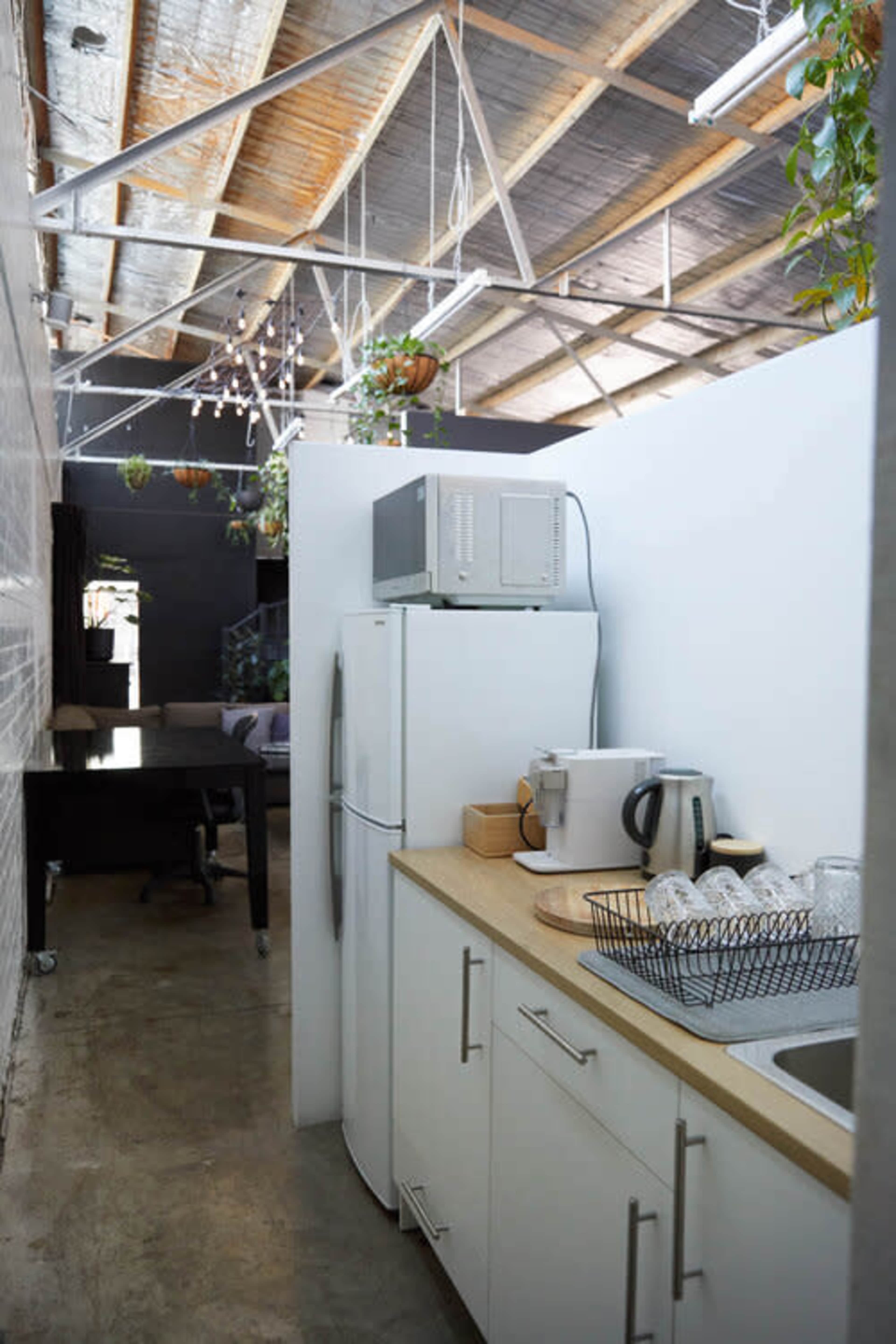 A modern kitchen features a refrigerator, microwave, and various kitchenware, leading into a spacious living area with exposed beams and greenery.