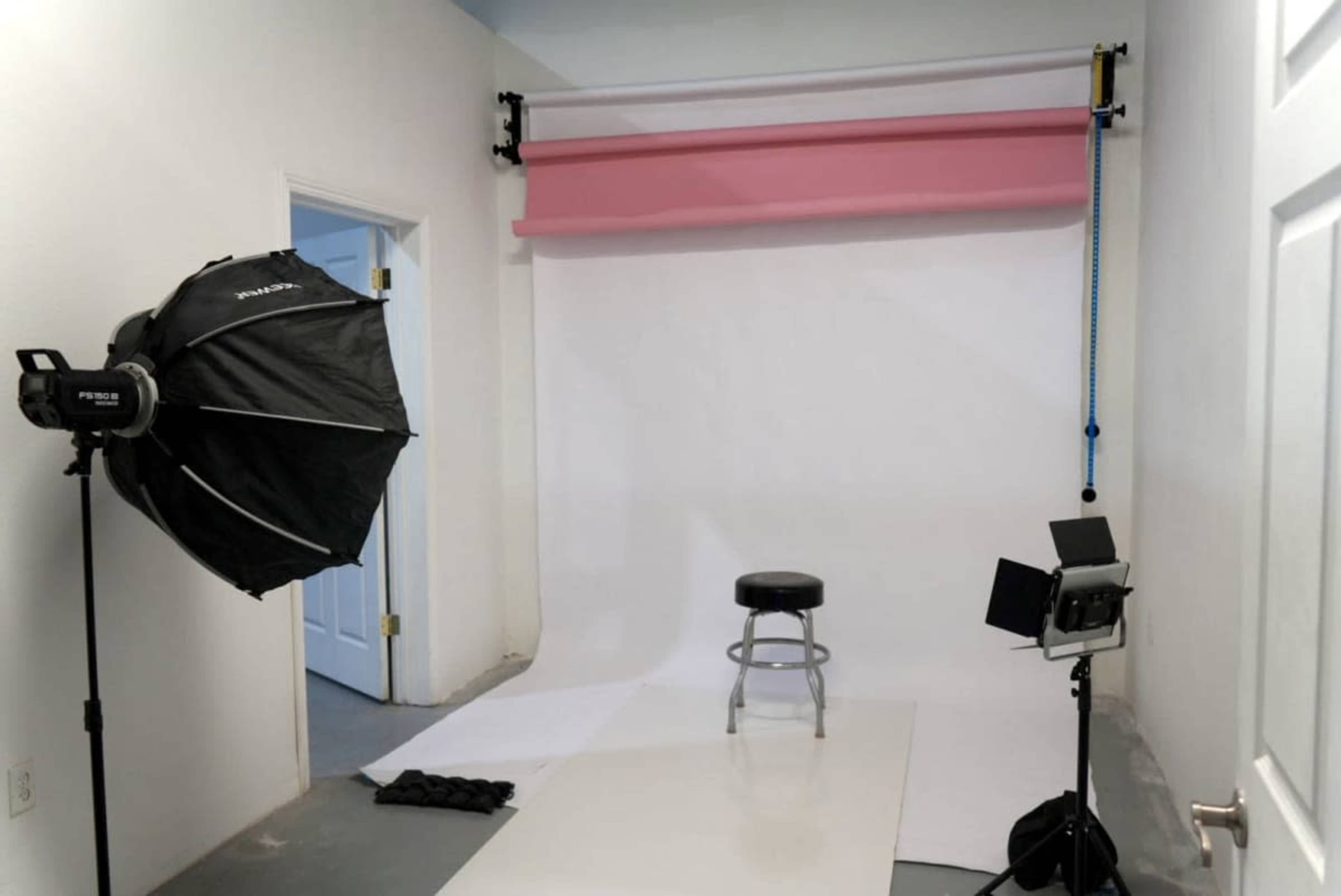 The image shows a photography studio with a white backdrop, a pink roll-up paper, a stool in the center, and two lighting setups on either side.