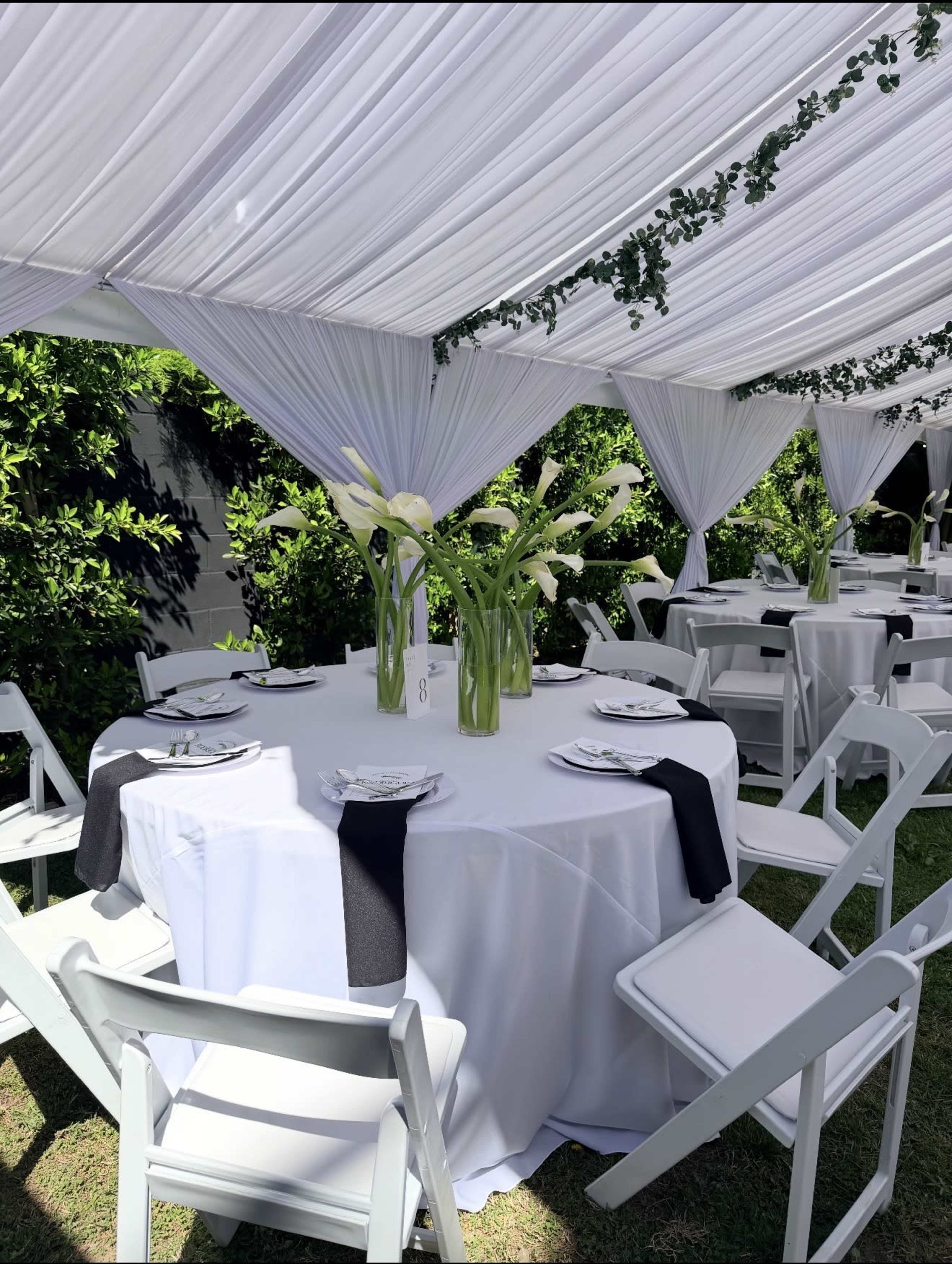 A round table is set under a white draped canopy, adorned with elegant floral centerpieces and neatly arranged place settings.