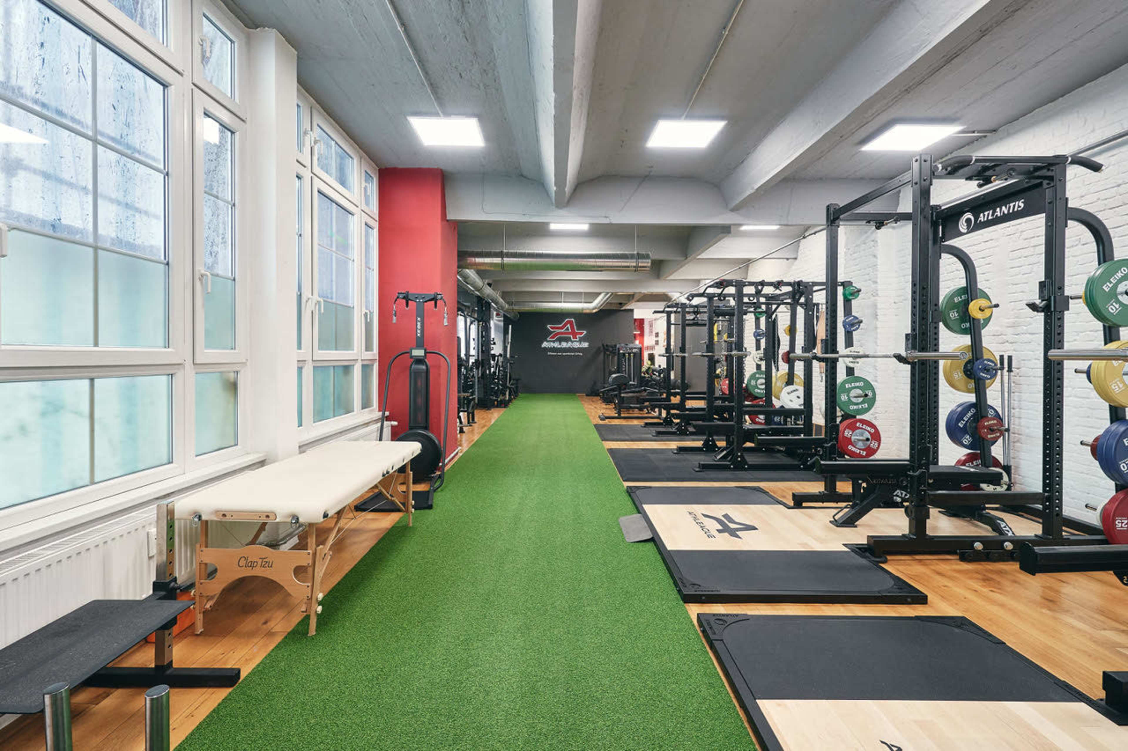 The image shows a spacious gym interior with weightlifting equipment, workout benches, and artificial turf flooring.