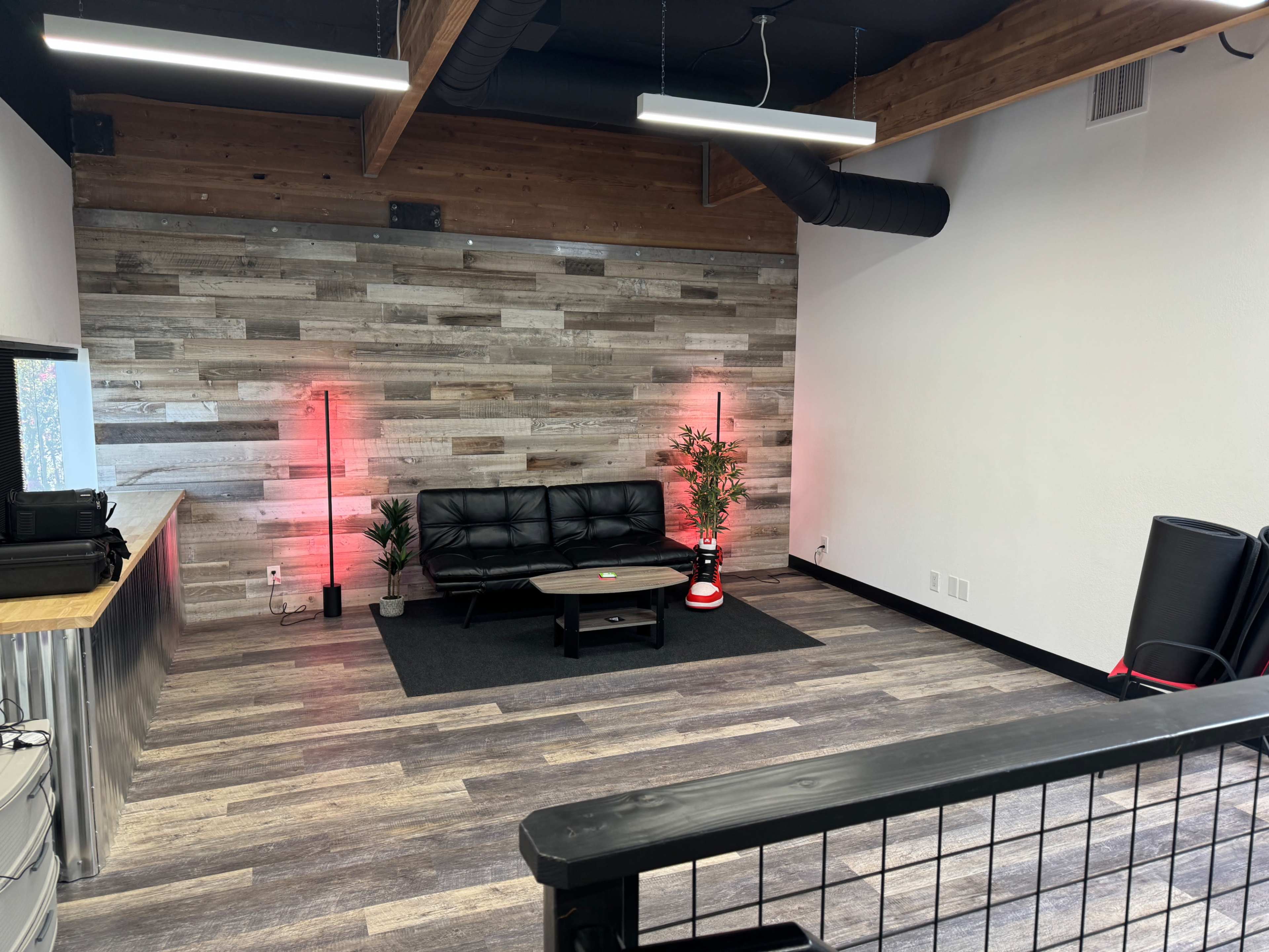A modern office space with a black couch, a wooden accent wall, and plants, illuminated by red light lamps.