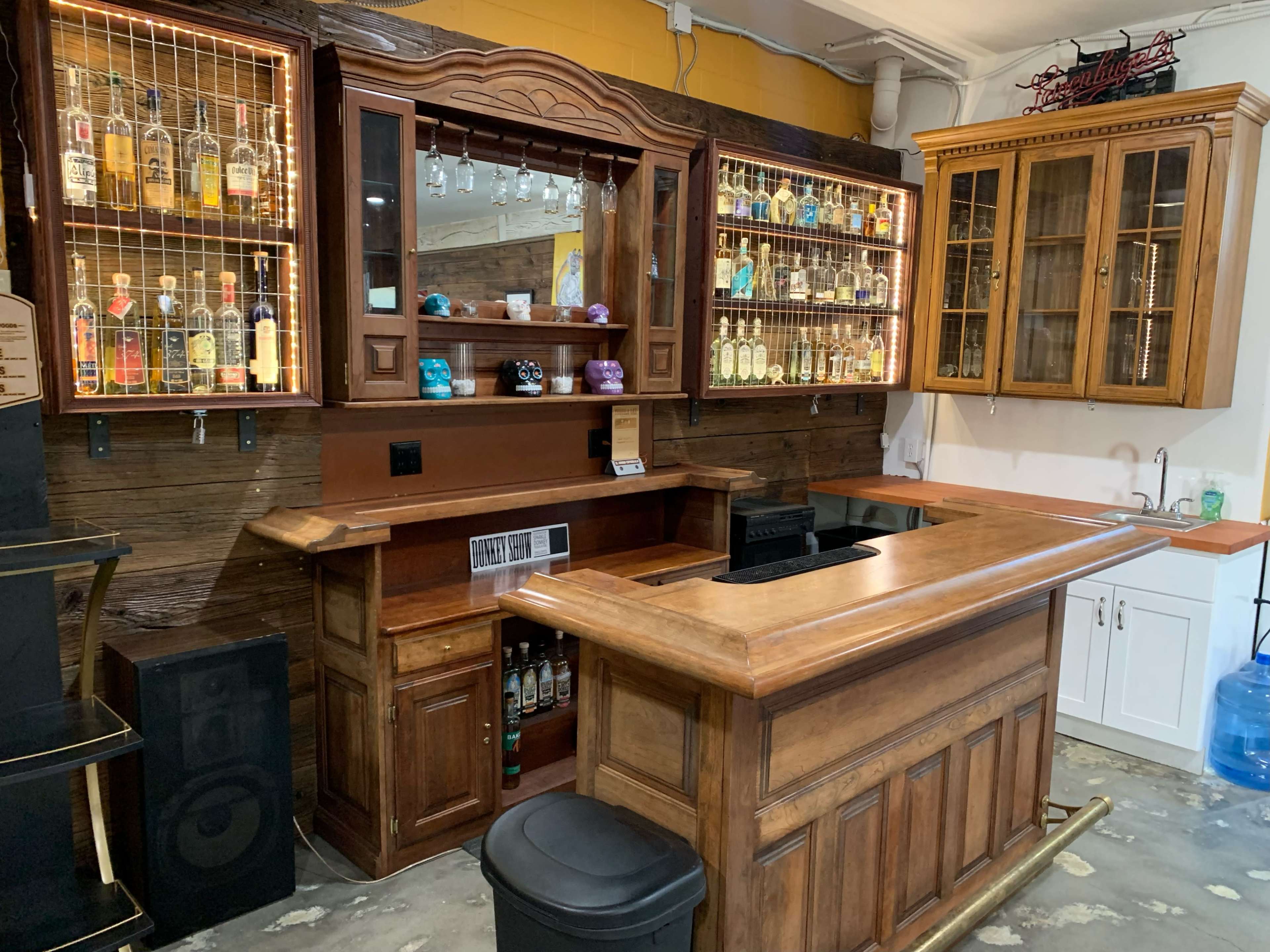 A wooden bar setup with an organized display of liquor bottles on shelves illuminated by lights.