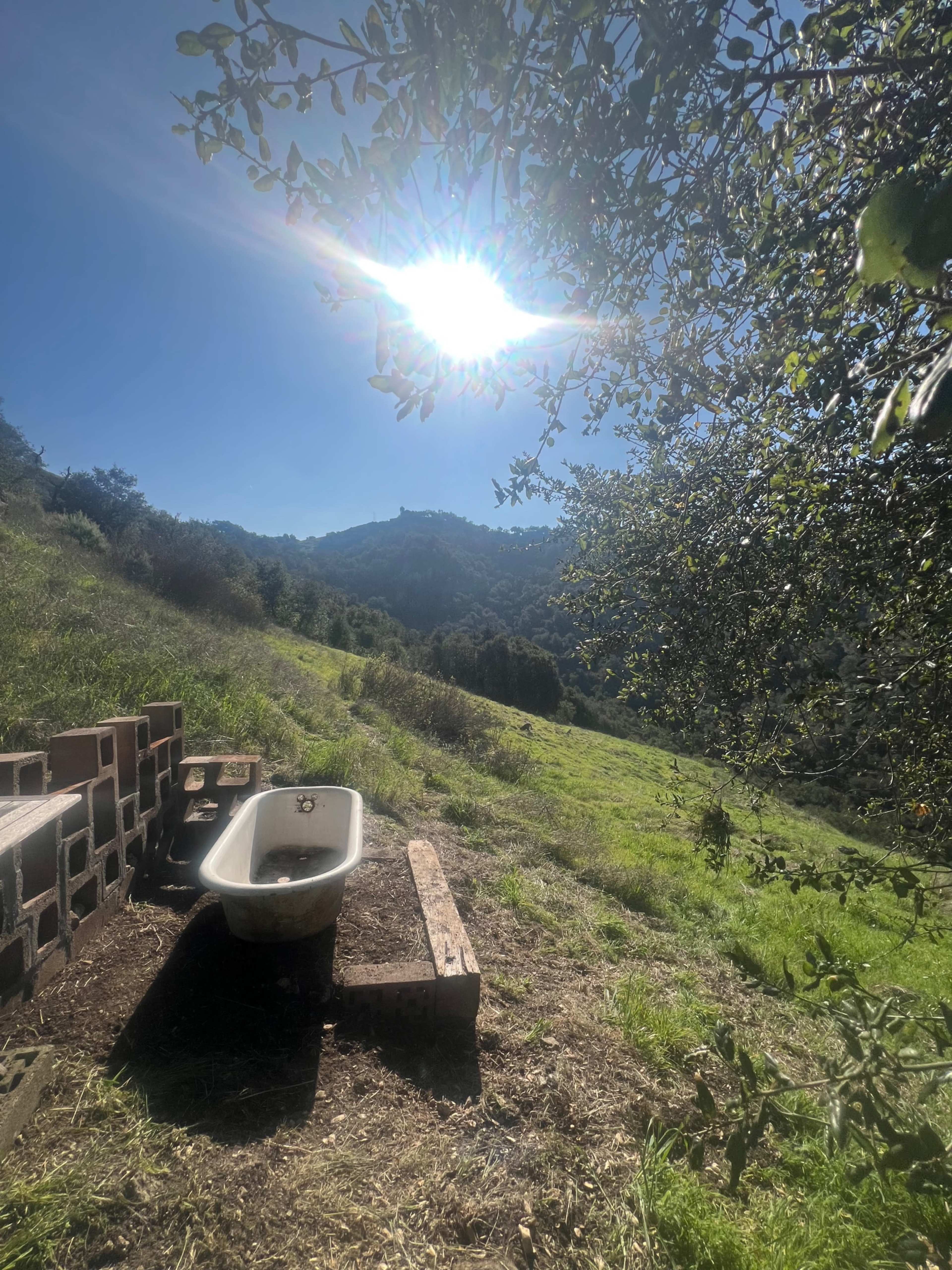 A vintage bathtub is placed on the edge of a grassy hillside, with sunlight gleaming above and mountains in the distance.