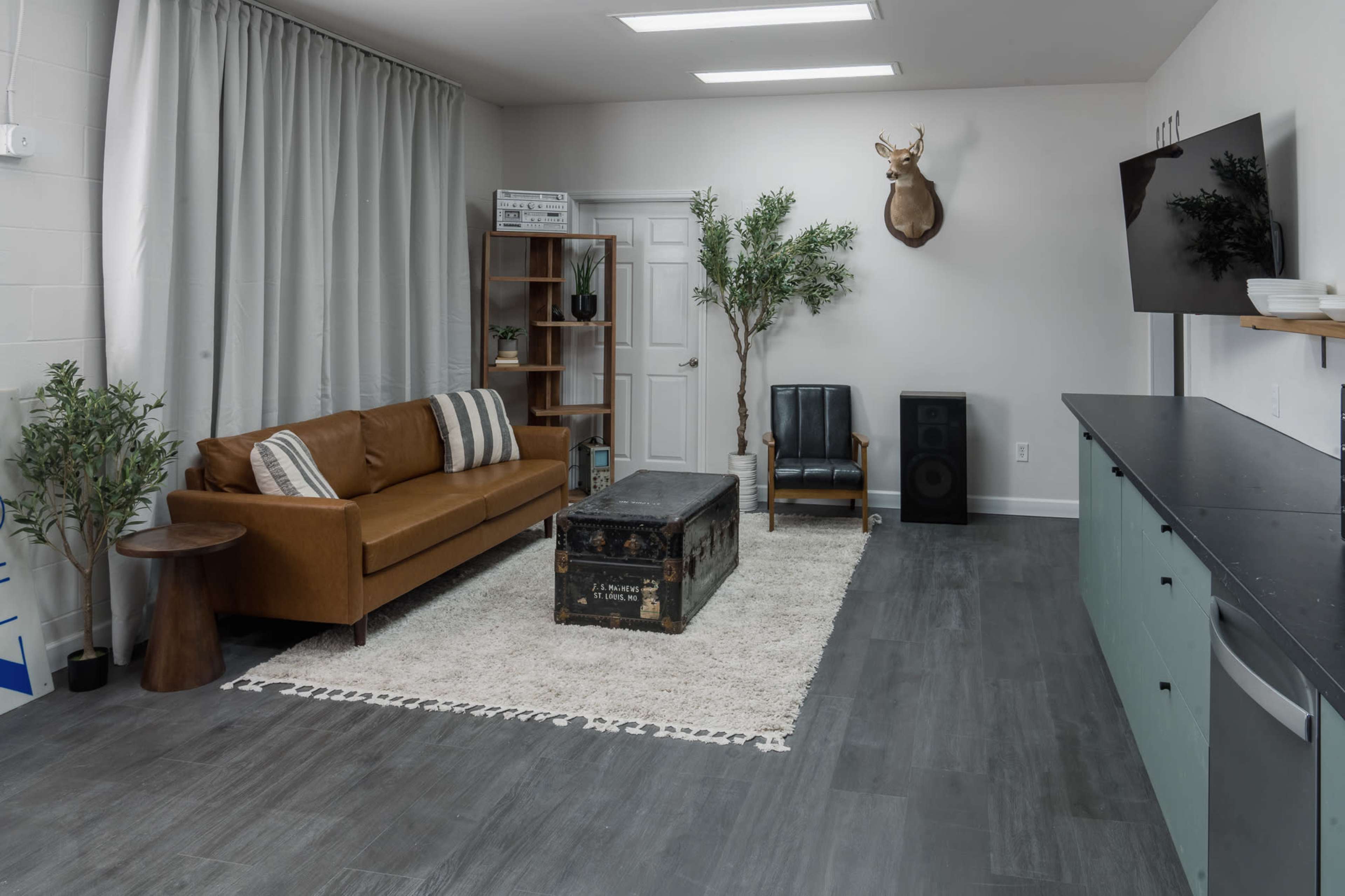 The image shows a modern living space featuring a brown leather sofa, a vintage trunk coffee table, and a deer head mounted on the wall, surrounded by greenery and a flat-screen TV.