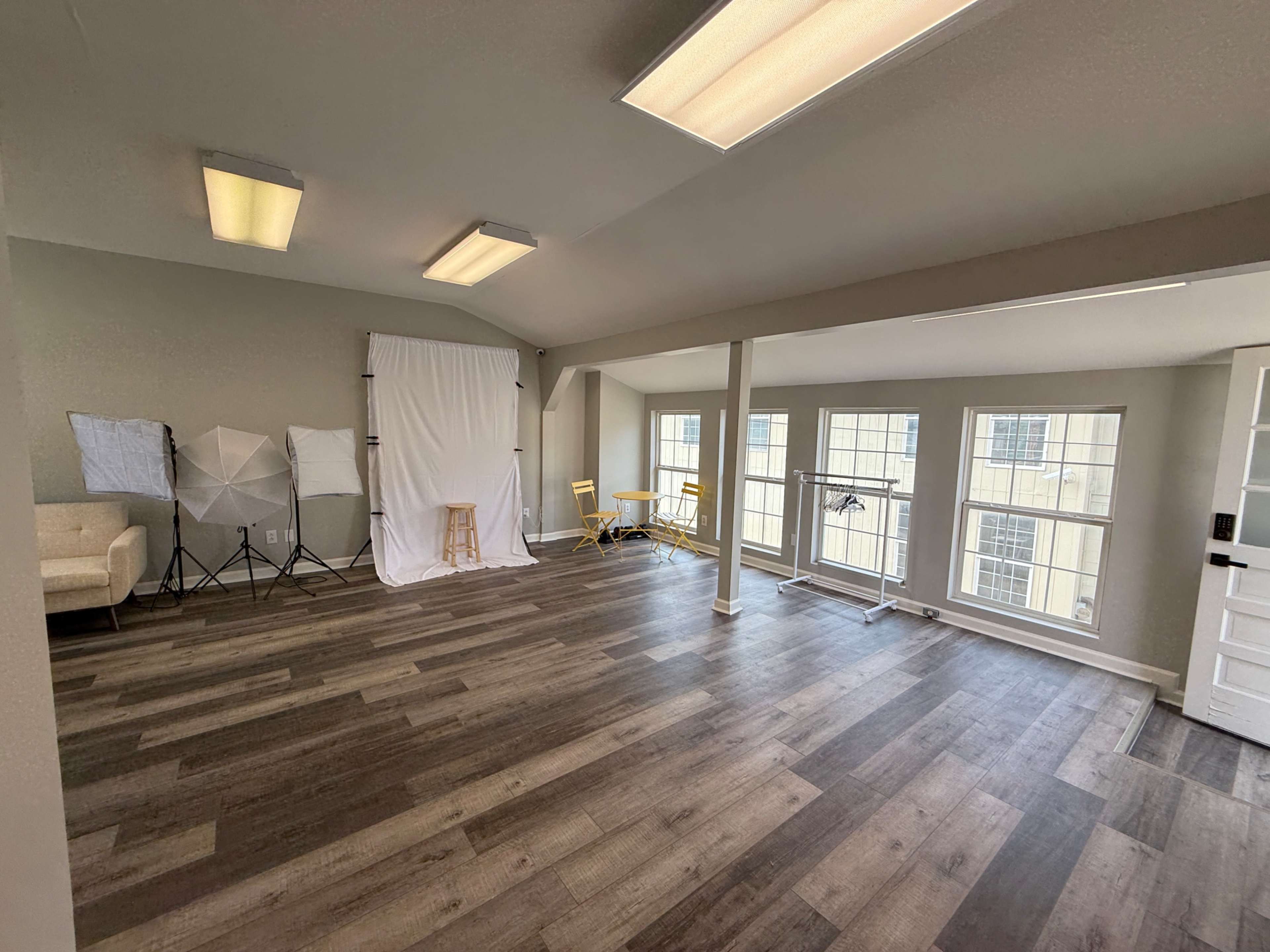 The image shows a spacious room with large windows, light wooden floors, and a photo backdrop setup including a chair and several light modifiers.