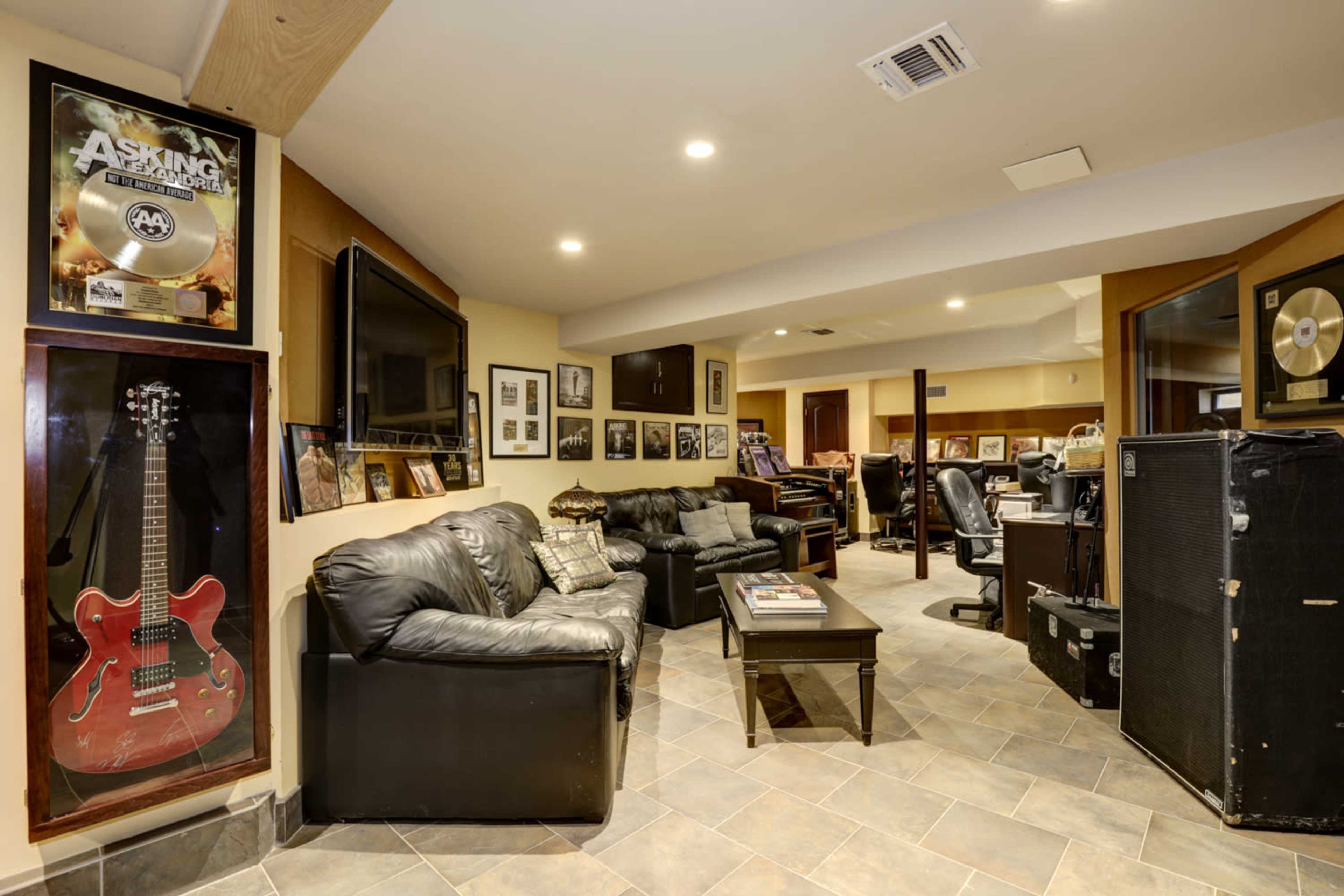 A basement lounge area featuring black leather sofas, a coffee table, framed music memorabilia, and electronic equipment.