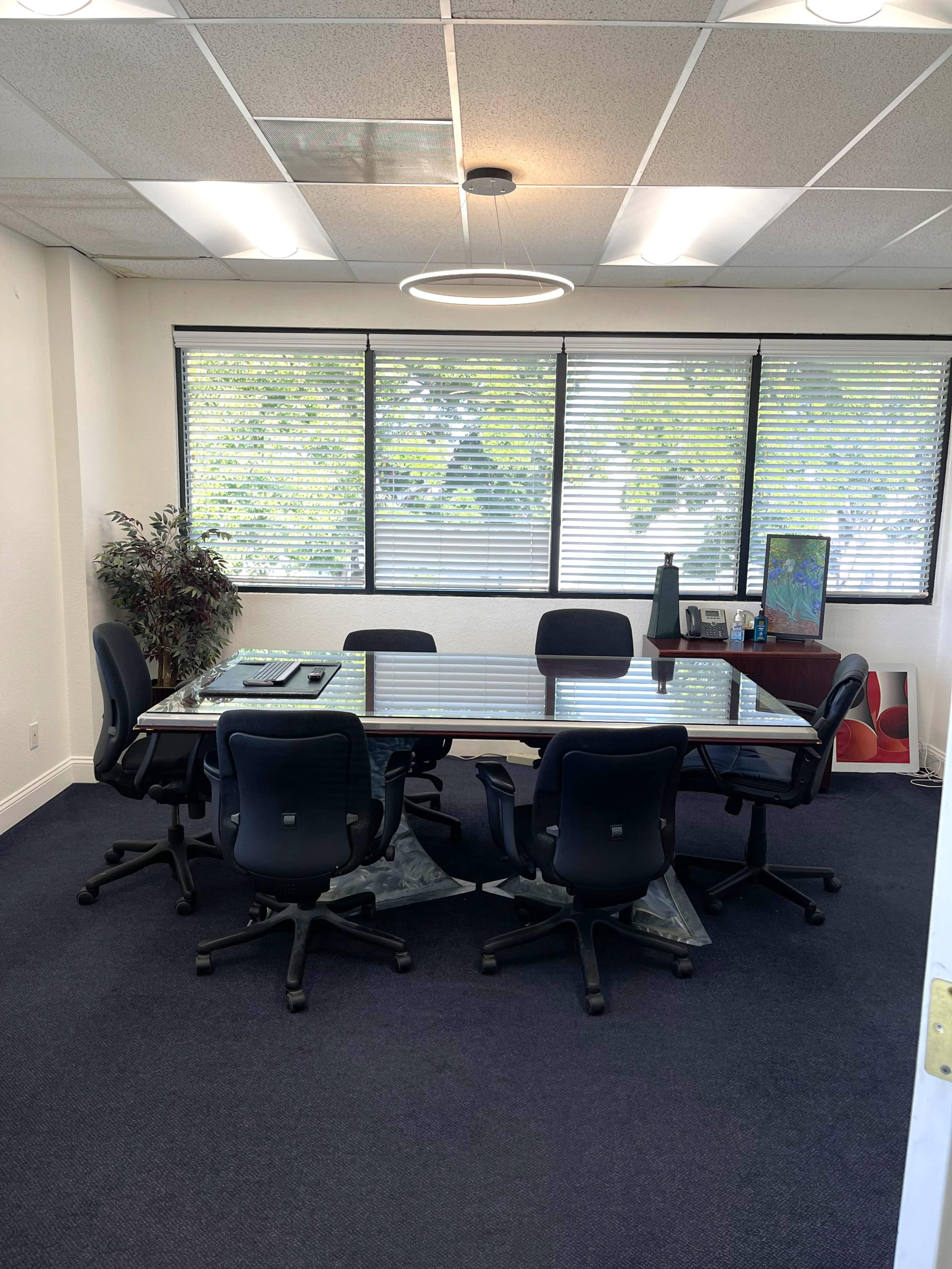 A conference room features a large glass table surrounded by six black ergonomic chairs, with large windows providing natural light.