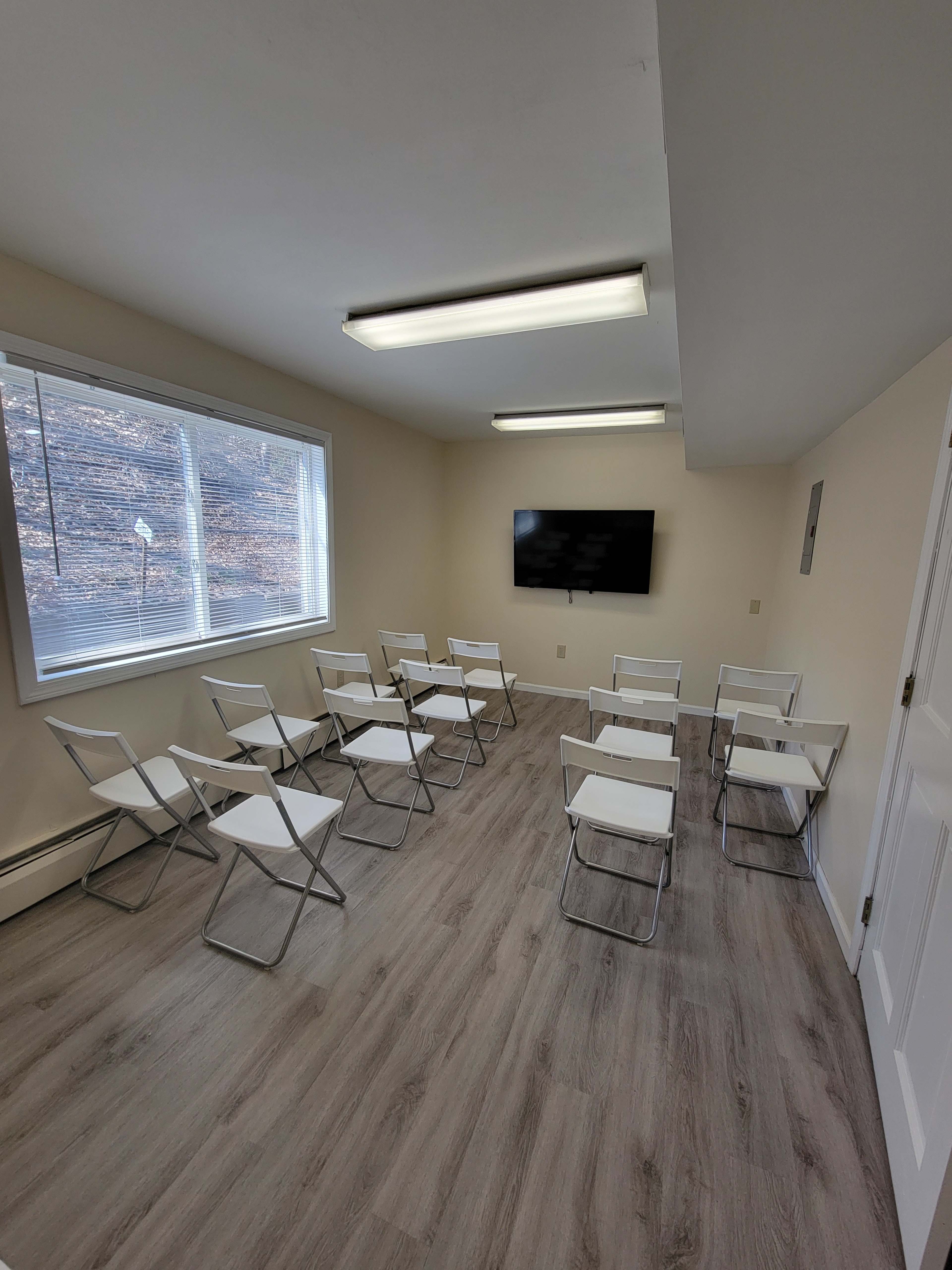 A small meeting room with white folding chairs arranged in rows facing a wall-mounted television and a large window providing natural light.