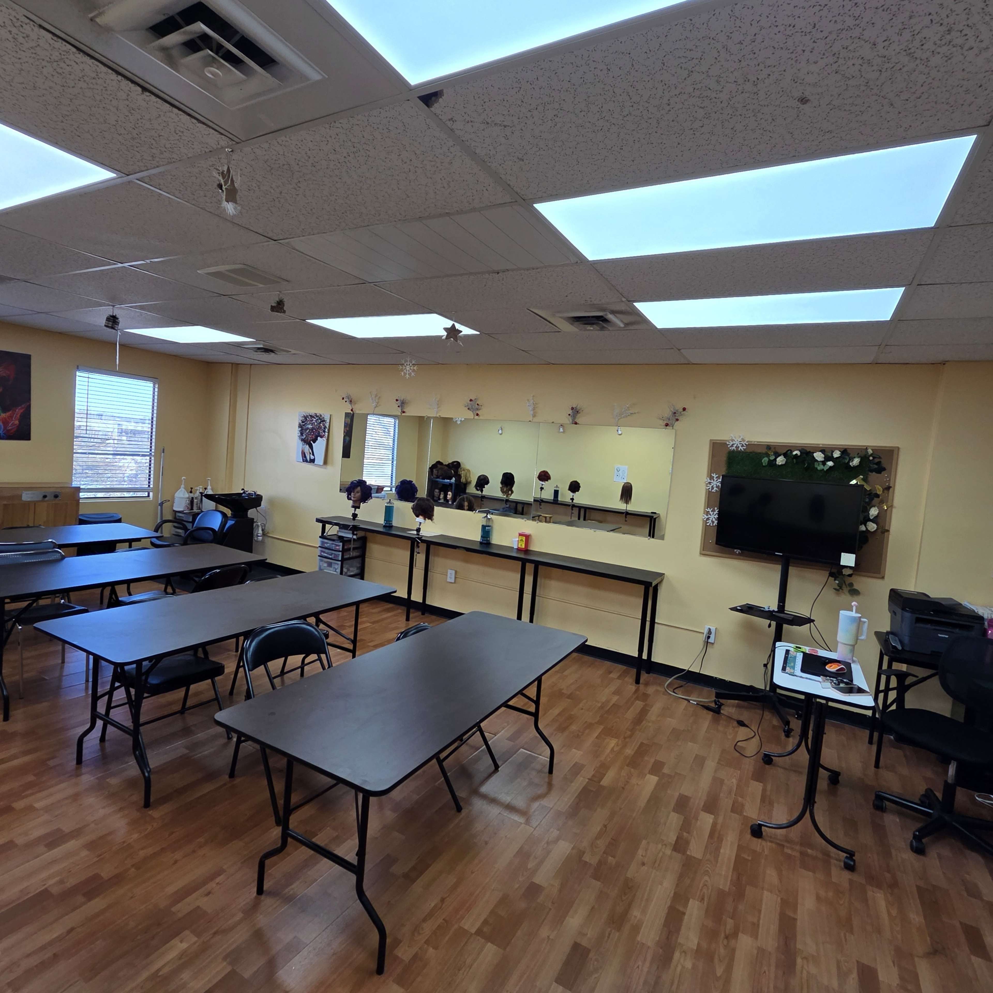 The image shows a classroom or workshop space with several tables arranged around the room, equipped with a television and mirrors on the wall.