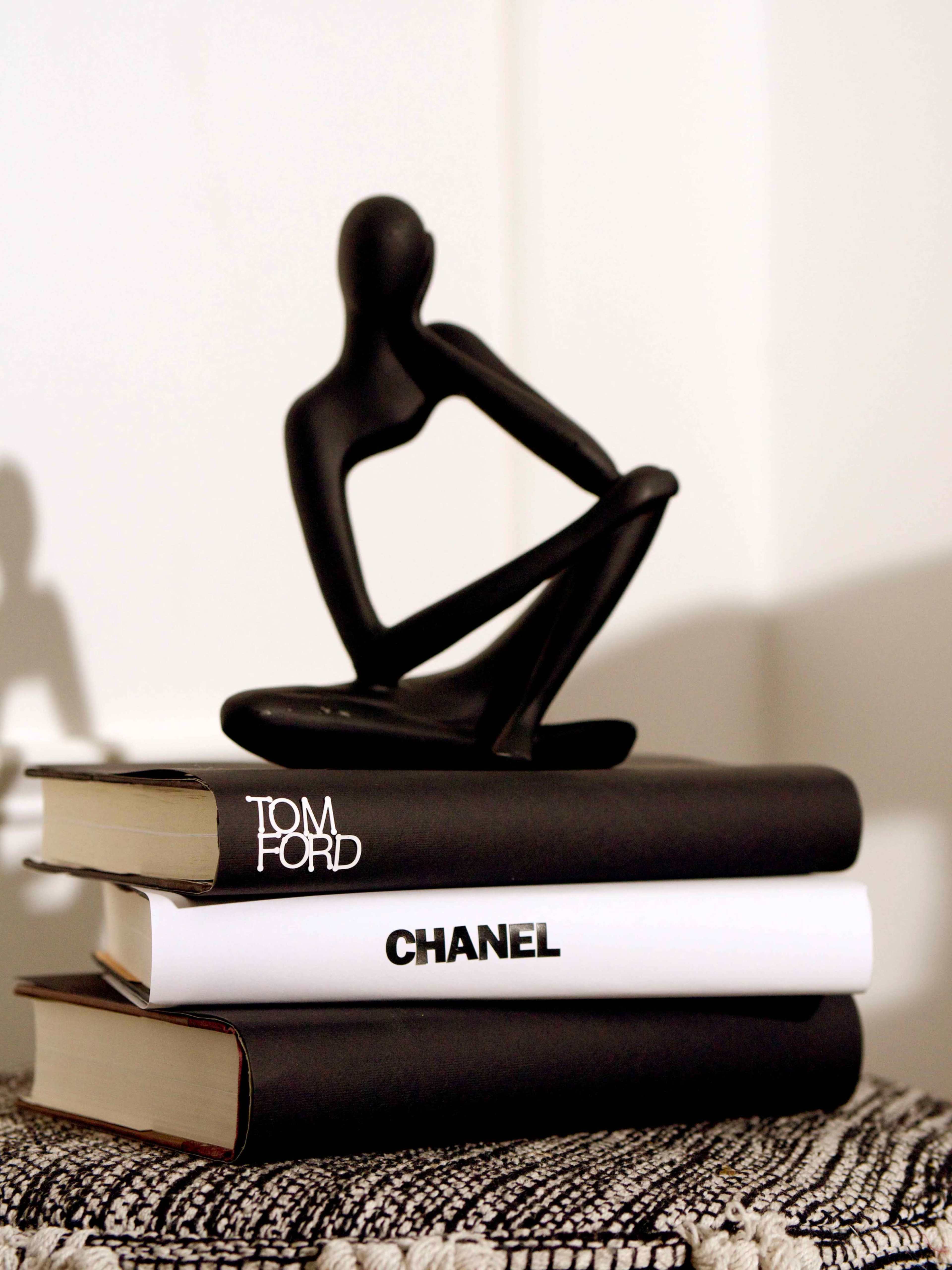 A black figurine sits atop a stack of three books, labeled "TOM FORD," "CHANEL," and a third book with a white cover.