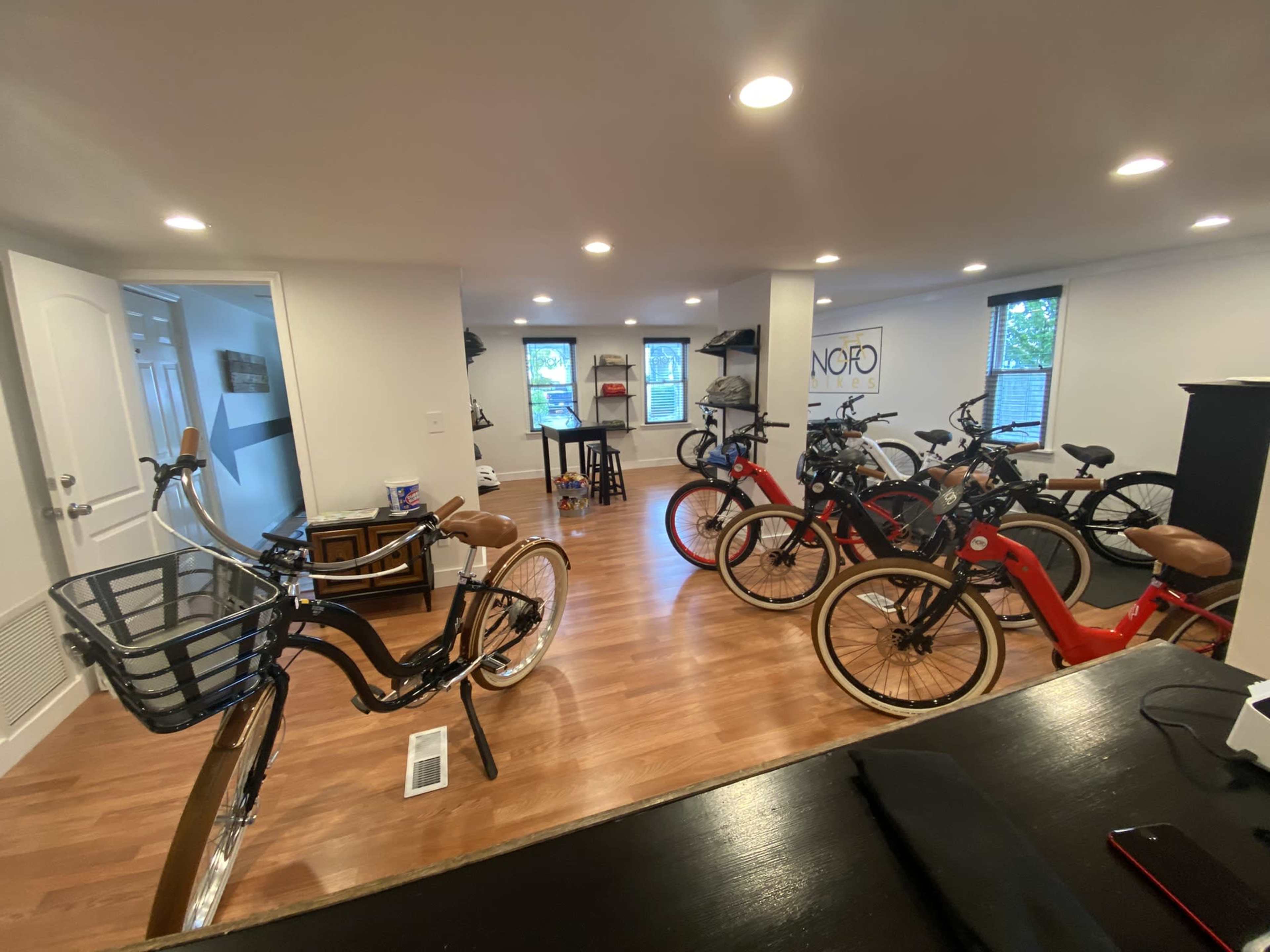 A spacious room filled with several bicycles, including traditional and electric models, arranged around wooden flooring and bright overhead lighting.
