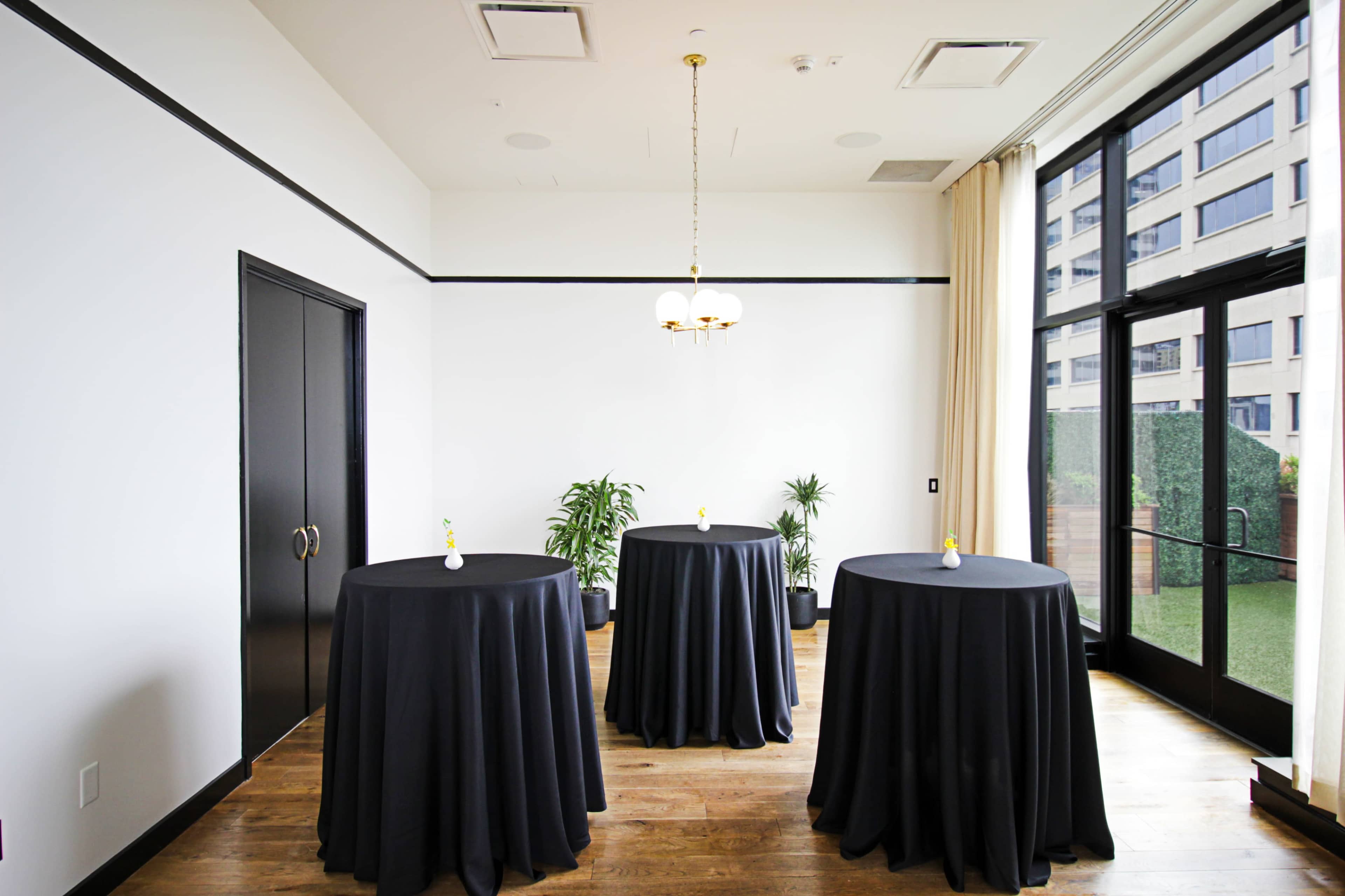 A bright room with three round tables covered in black tablecloths, arranged near large windows overlooking a green area.