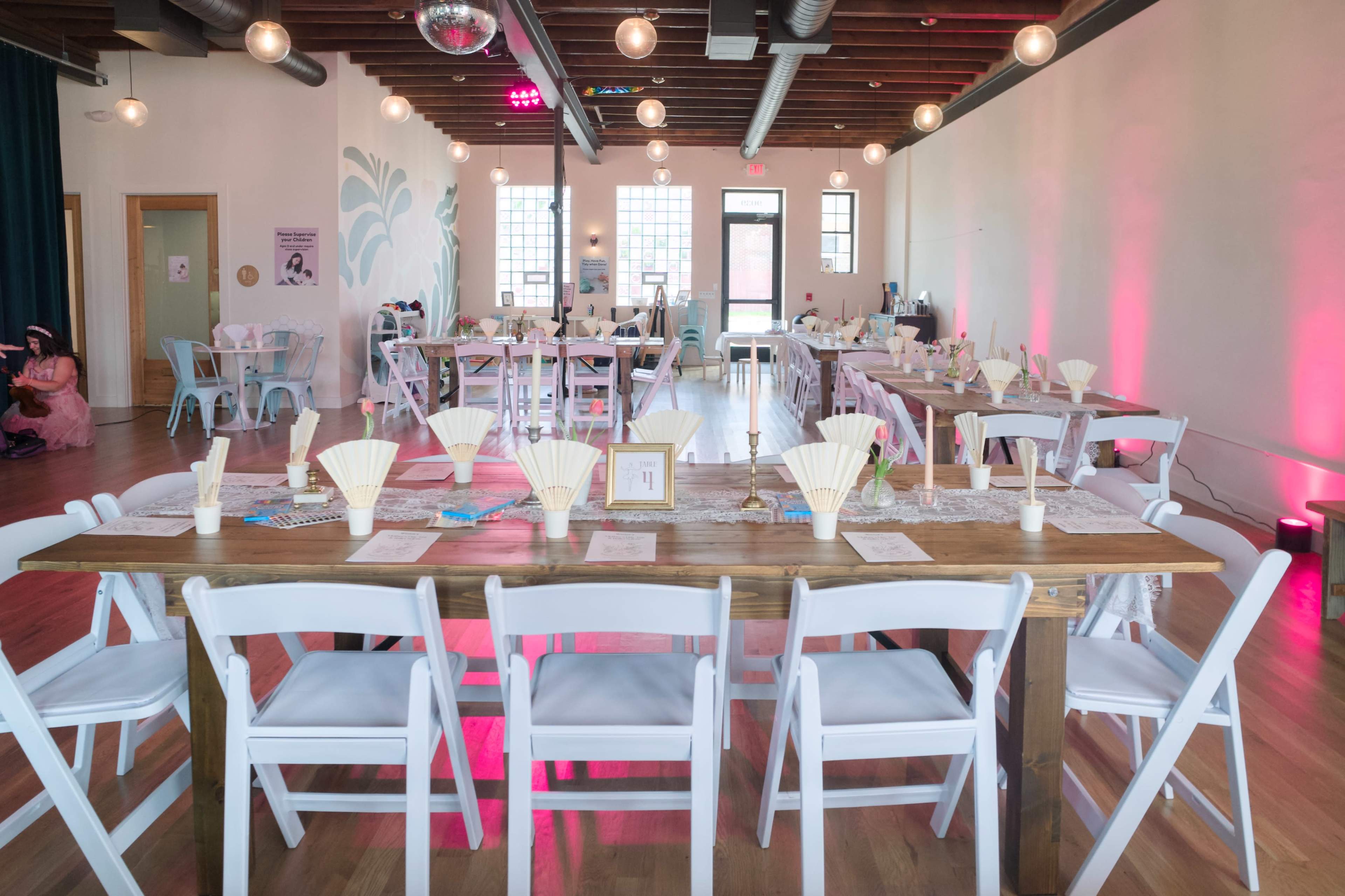 A spacious event room features several tables set with decorative fans, candles, and place settings, illuminated by hanging lights.
