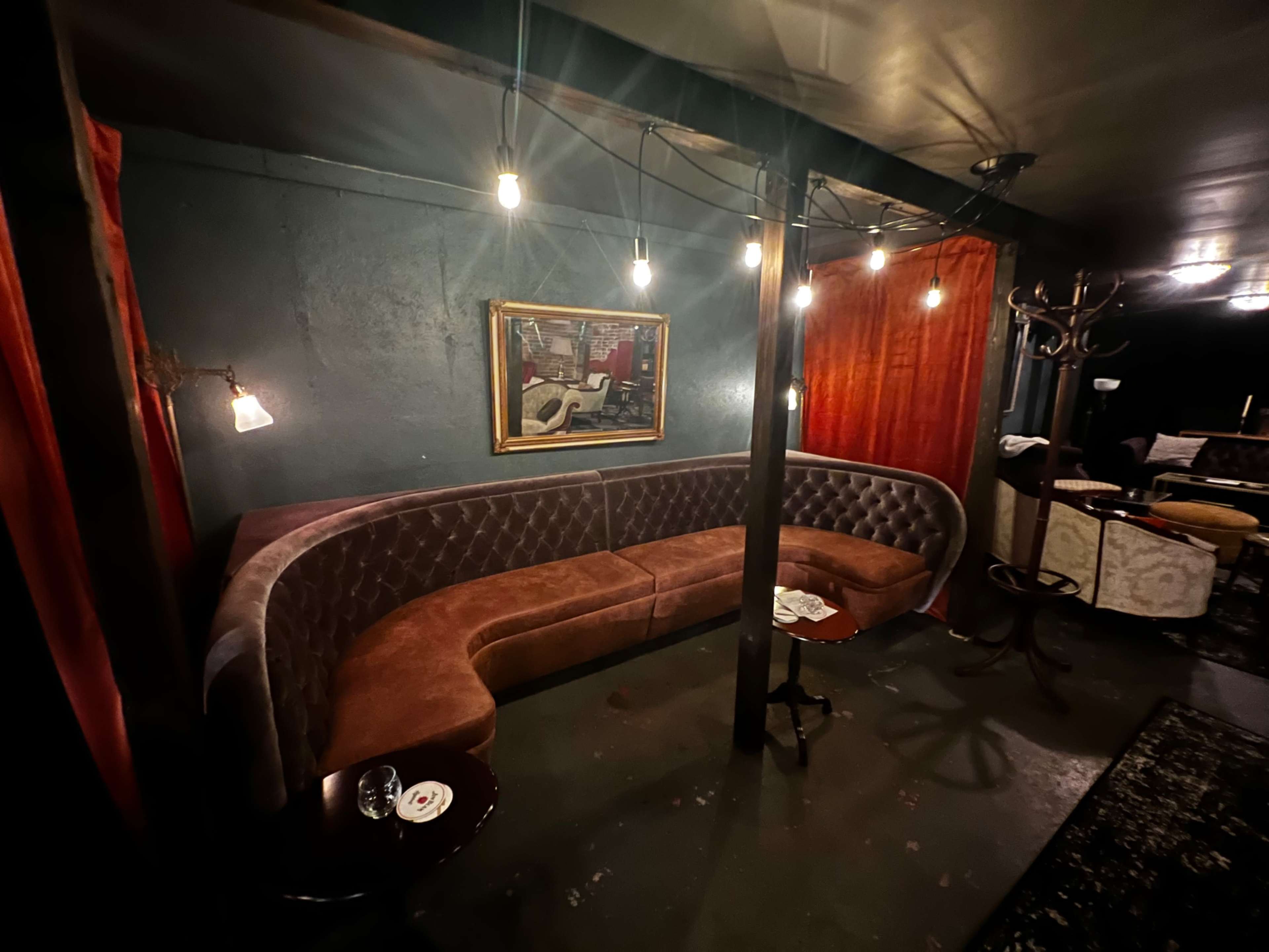 A dimly lit lounge area with a curved brown velvet couch, a small round table, and hanging light bulbs overhead.