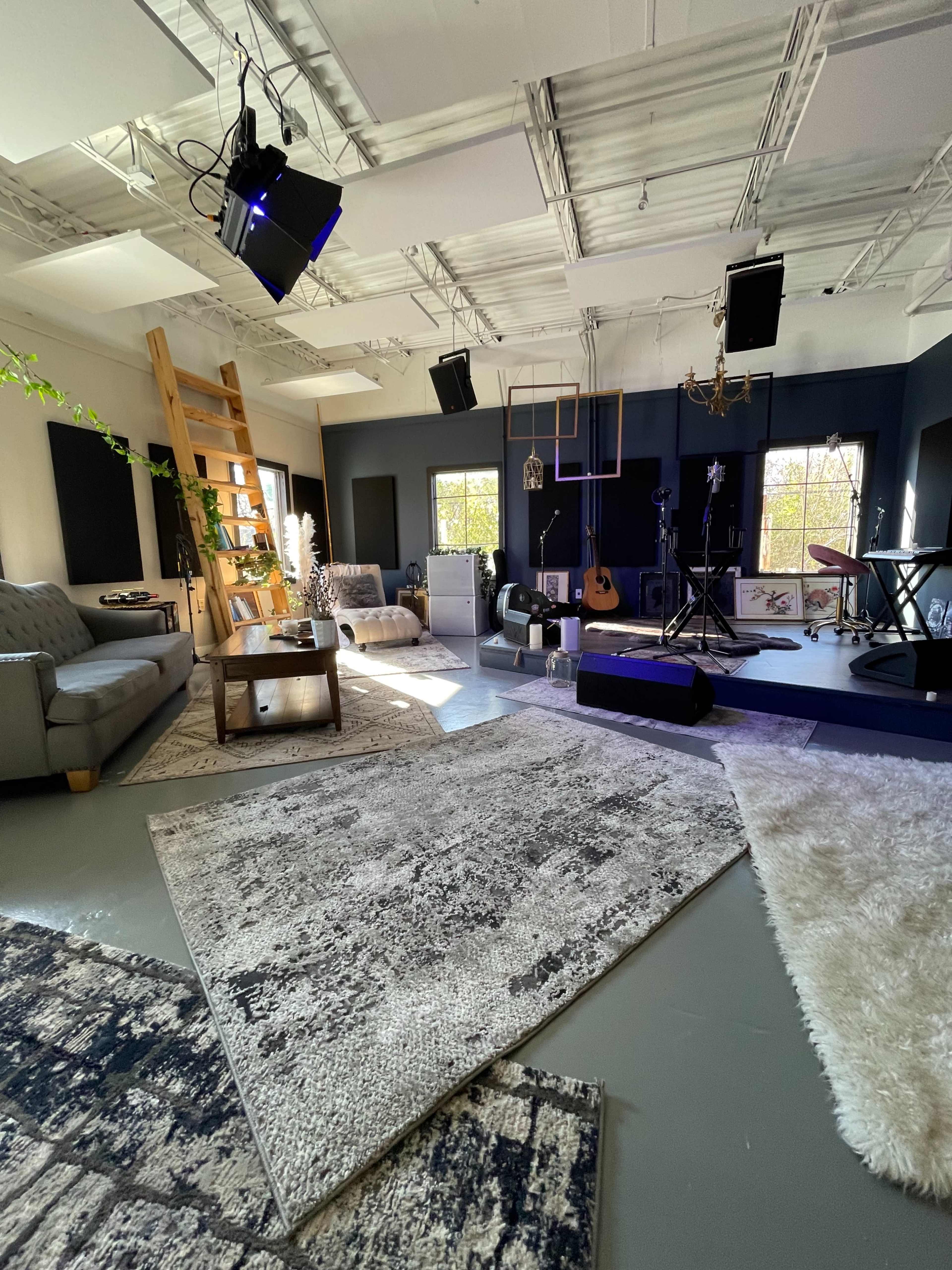 A well-lit studio space featuring comfortable seating, a wooden shelf, musical instruments, and area rugs.