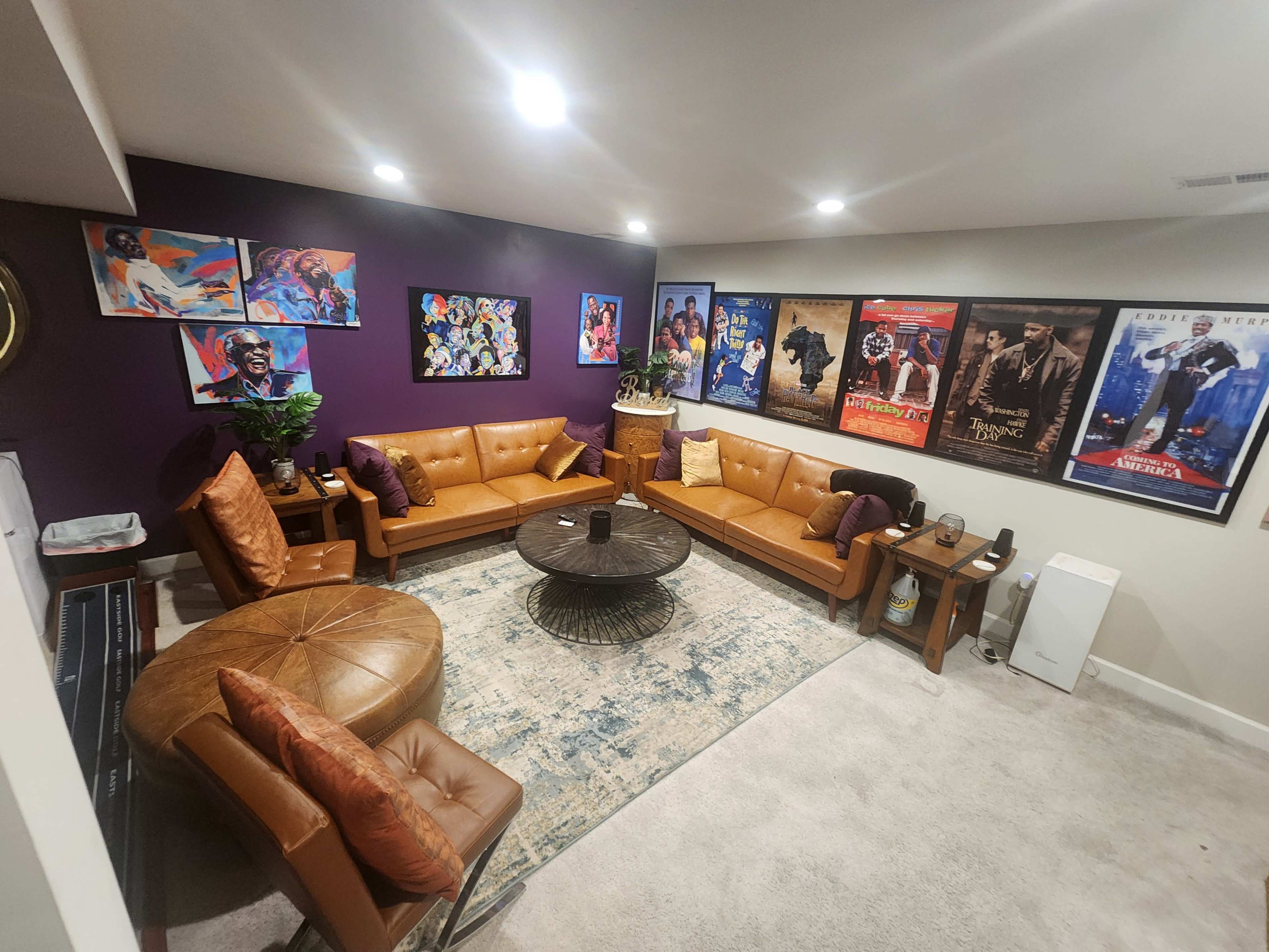 The image shows a cozy living room with brown leather sofas arranged around a circular black coffee table, surrounded by colorful framed movie posters on the walls.