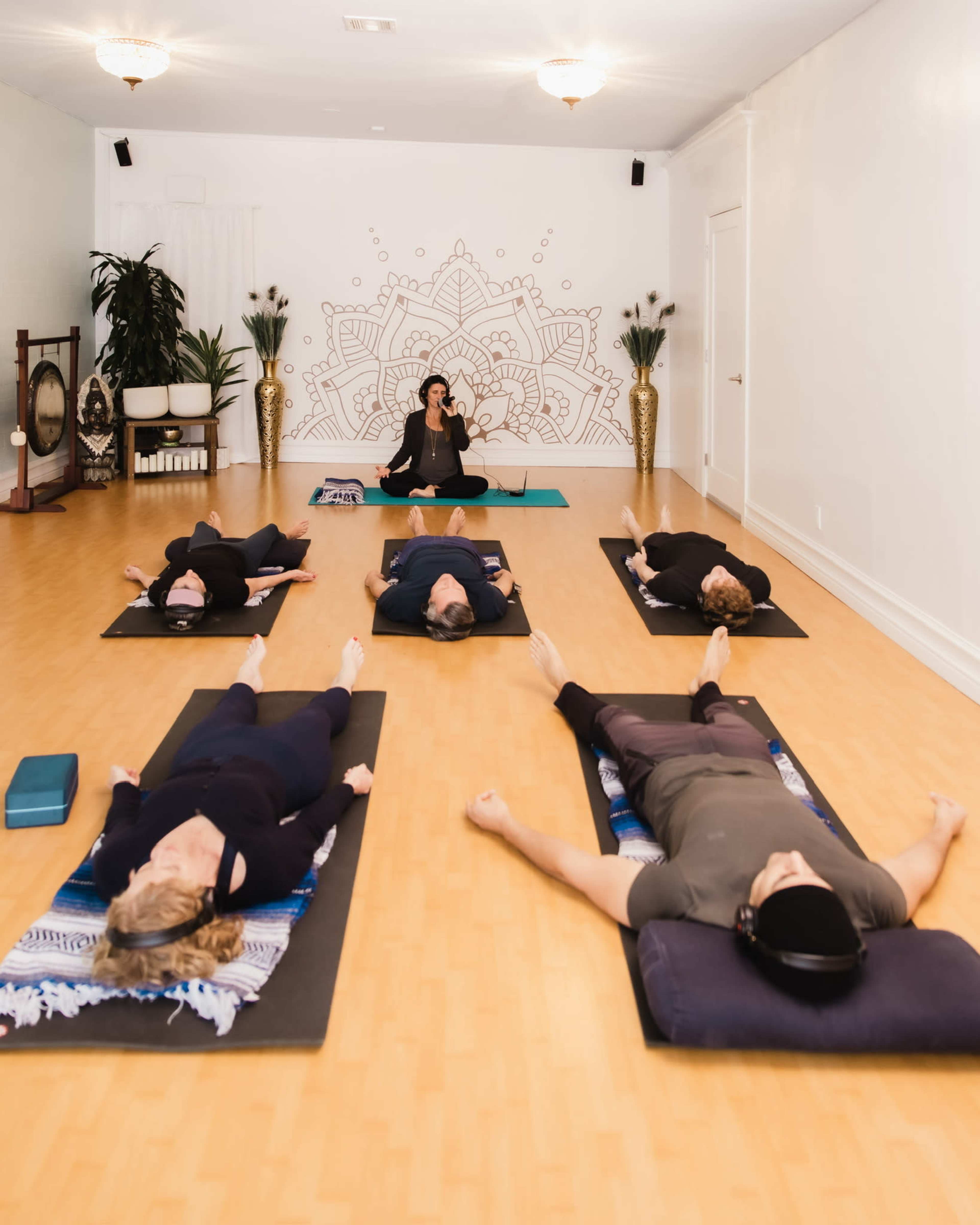 A yoga studio features several participants lying on mats in a relaxation pose while an instructor sits at the front, leading the session.