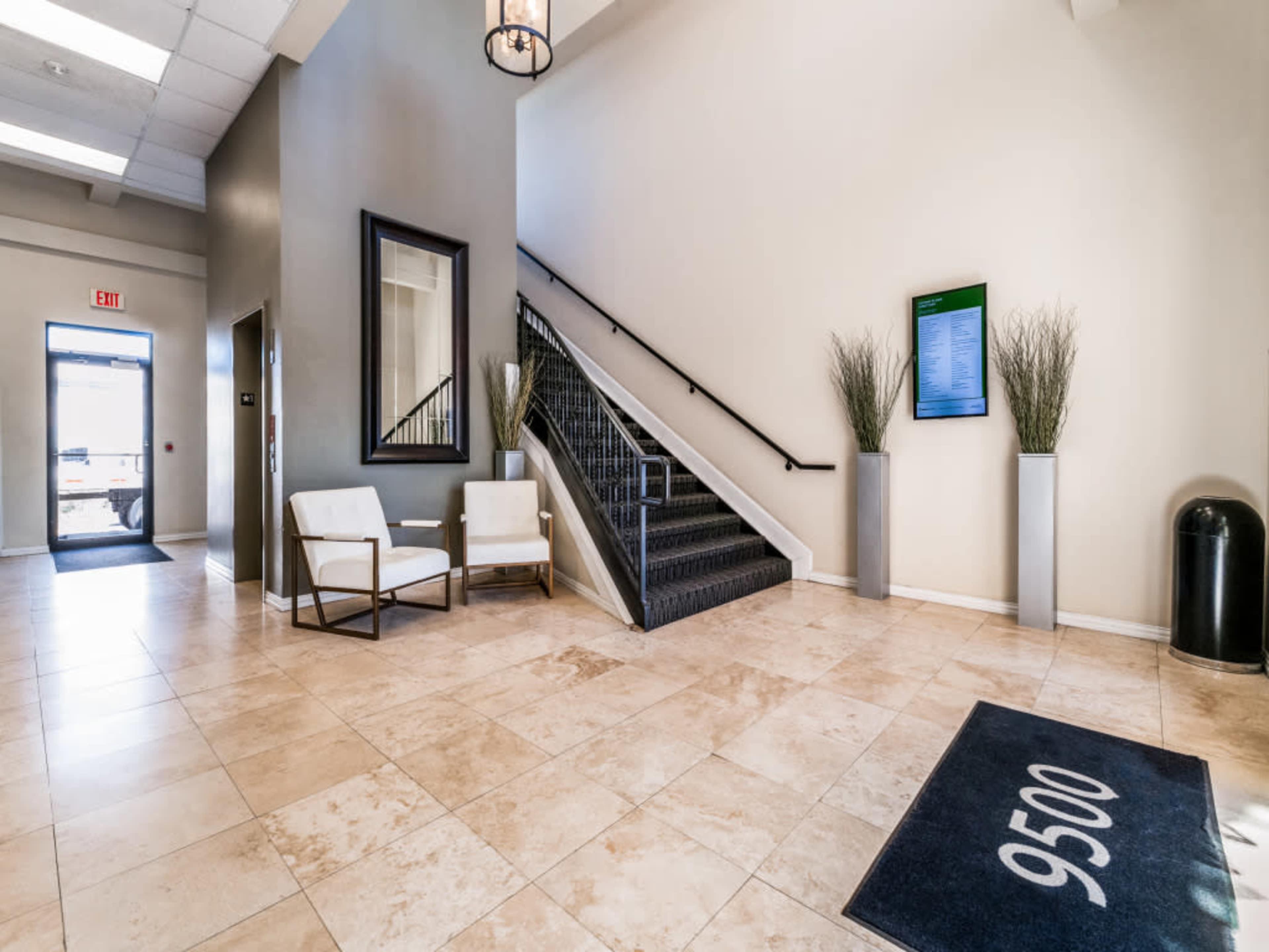 The image shows a spacious lobby area with a staircase, two white chairs, a tall plant in each corner, and a directory display on the wall.