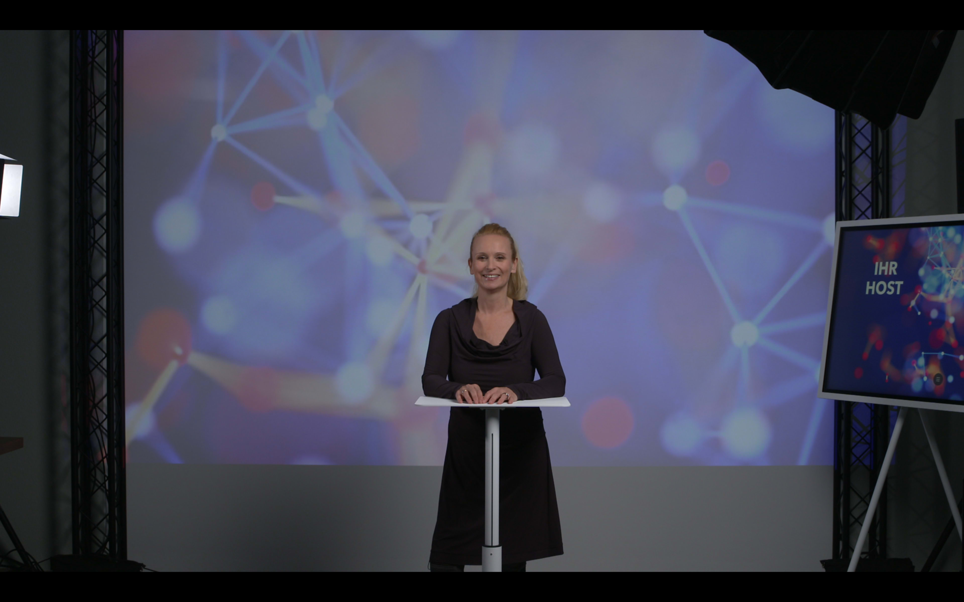 A woman stands behind a podium in front of a colorful, abstract background featuring interconnected lines and dots.